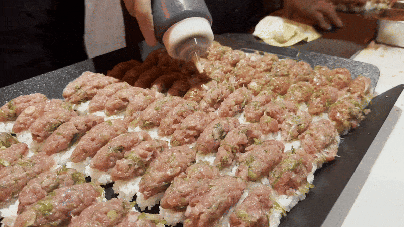 A person squeezes a dark brown sauce over a tray of formed tataki fillings.