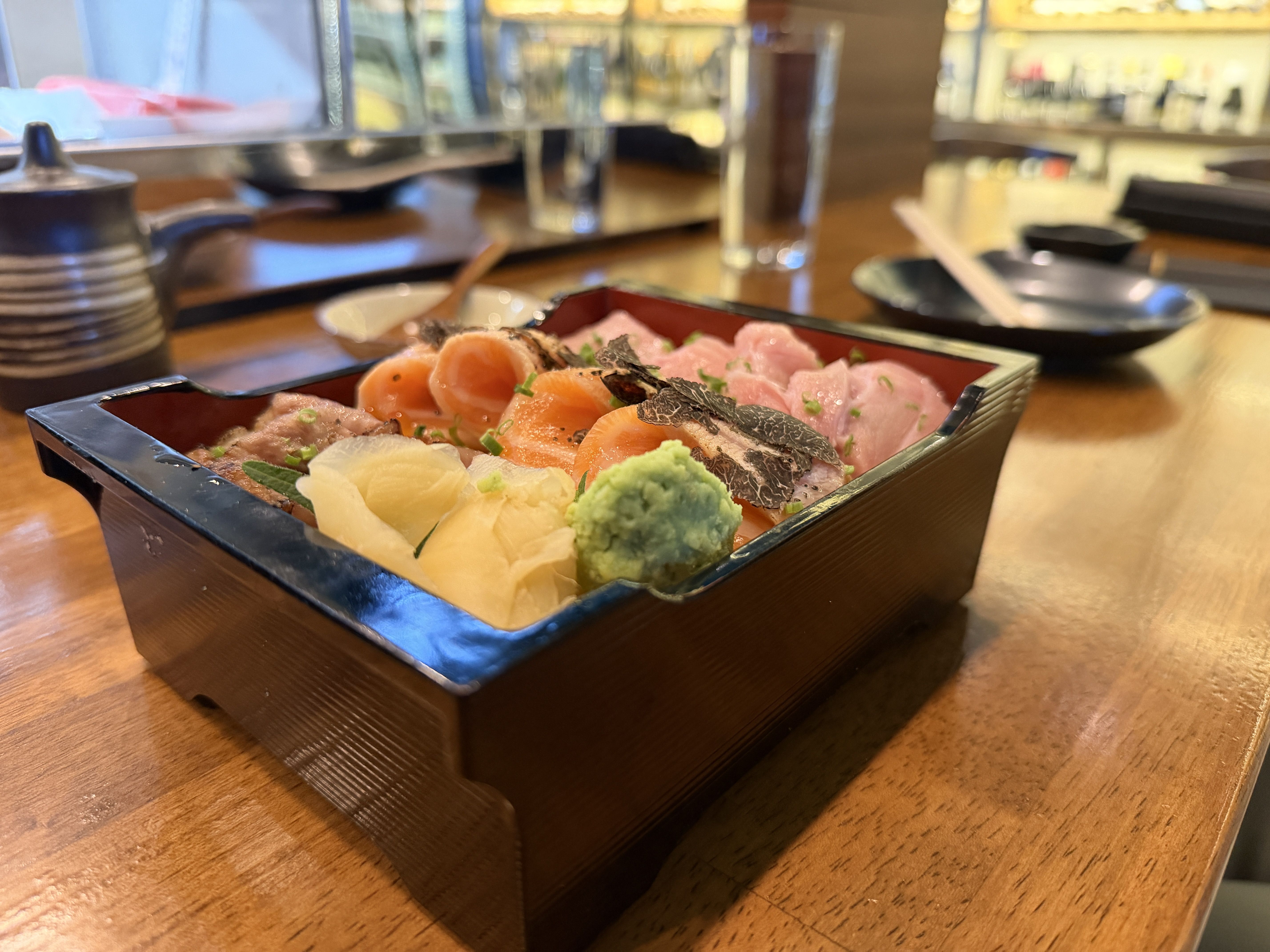 Black lacquered bento box with assorted sashimi including pink tuna, orange salmon with black truffle, green wasabi, and pickled ginger on a wooden table in a sushi restaurant.