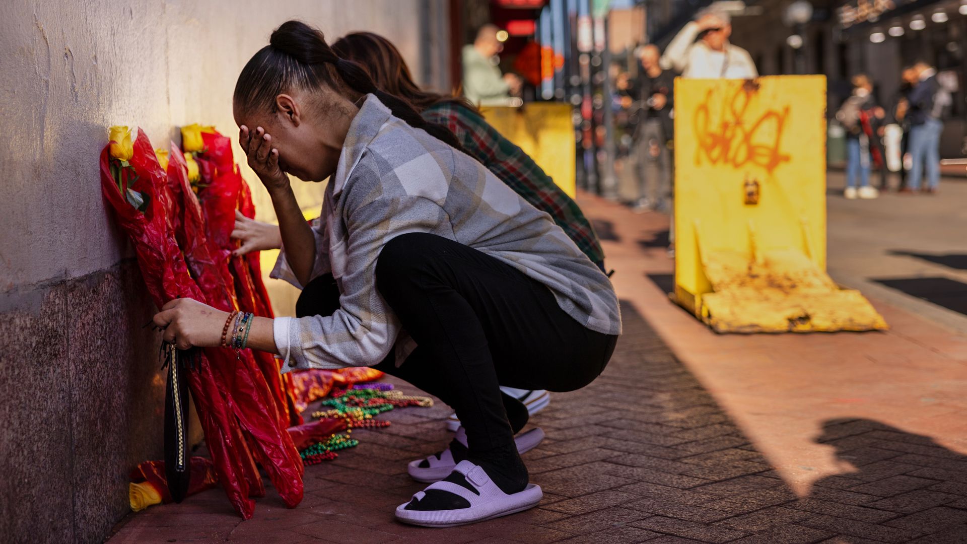 A person kneels down and mourns next to a row of yellow roses wrapped in red tissue paper. Behind them are two bright yellow sidewalk blockades.