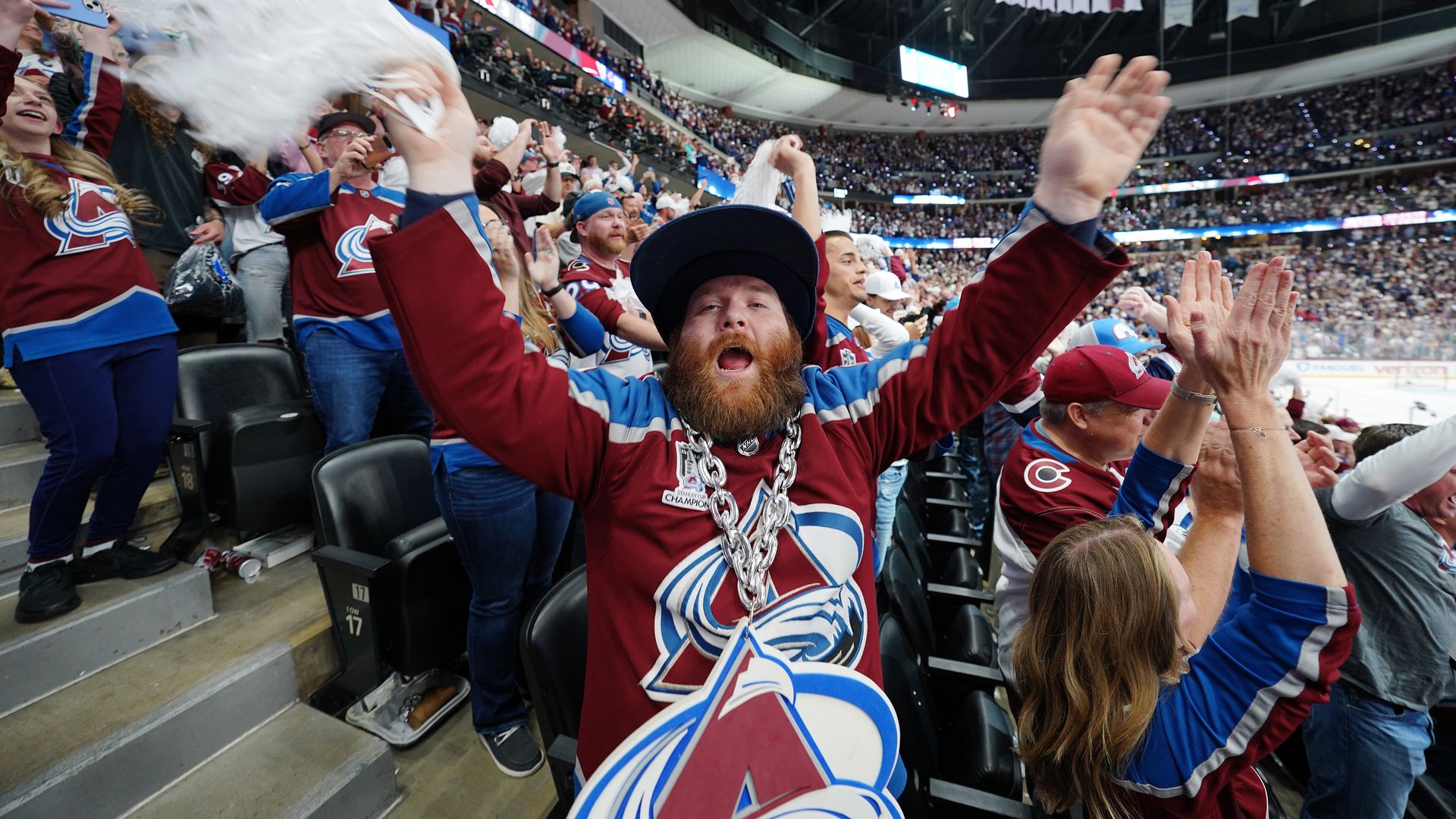 Fans fill a hockey arena, mostly wearing maroon, blue, and white Avalanche jerseys, cheering loudly. In the foreground a bearded man with a cap and a chunky silver chain raises his arms amid white scarves.