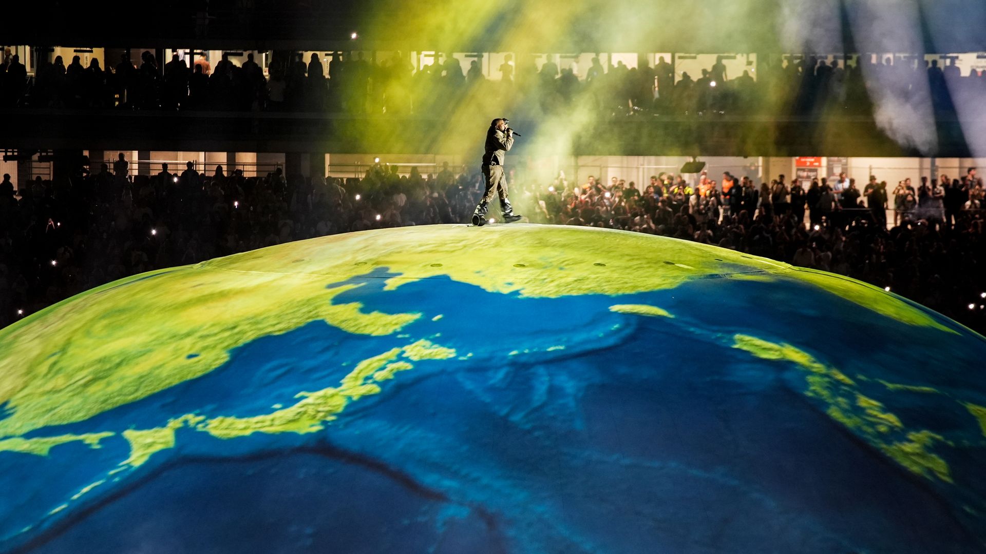 Ye holds a microphone to his face as he's viewed from a distance performing inside a stadium. The stage he walks along is spherical and designed like a globe.