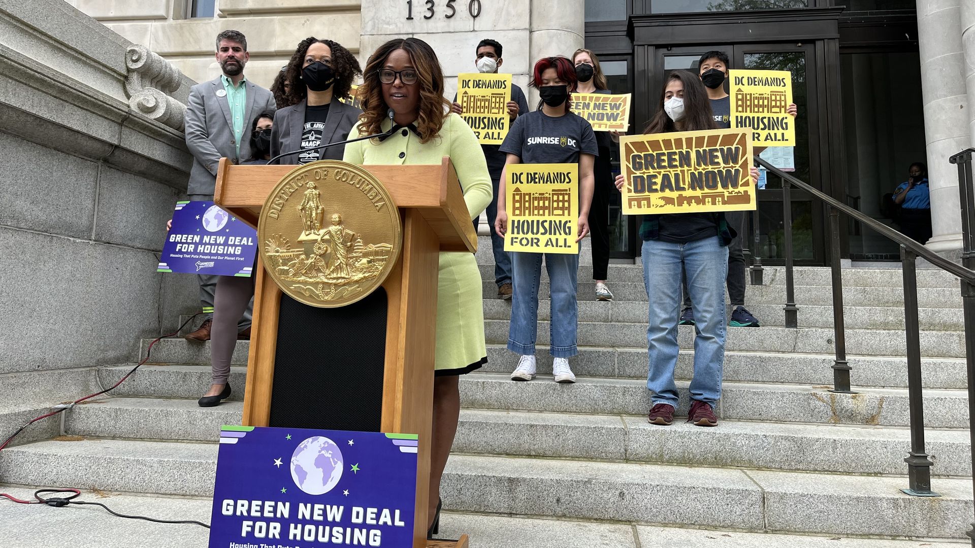 Janeese Lewis George stands behind a podium on the steps of a government building with supporters behind her holding signs for Green New Deal