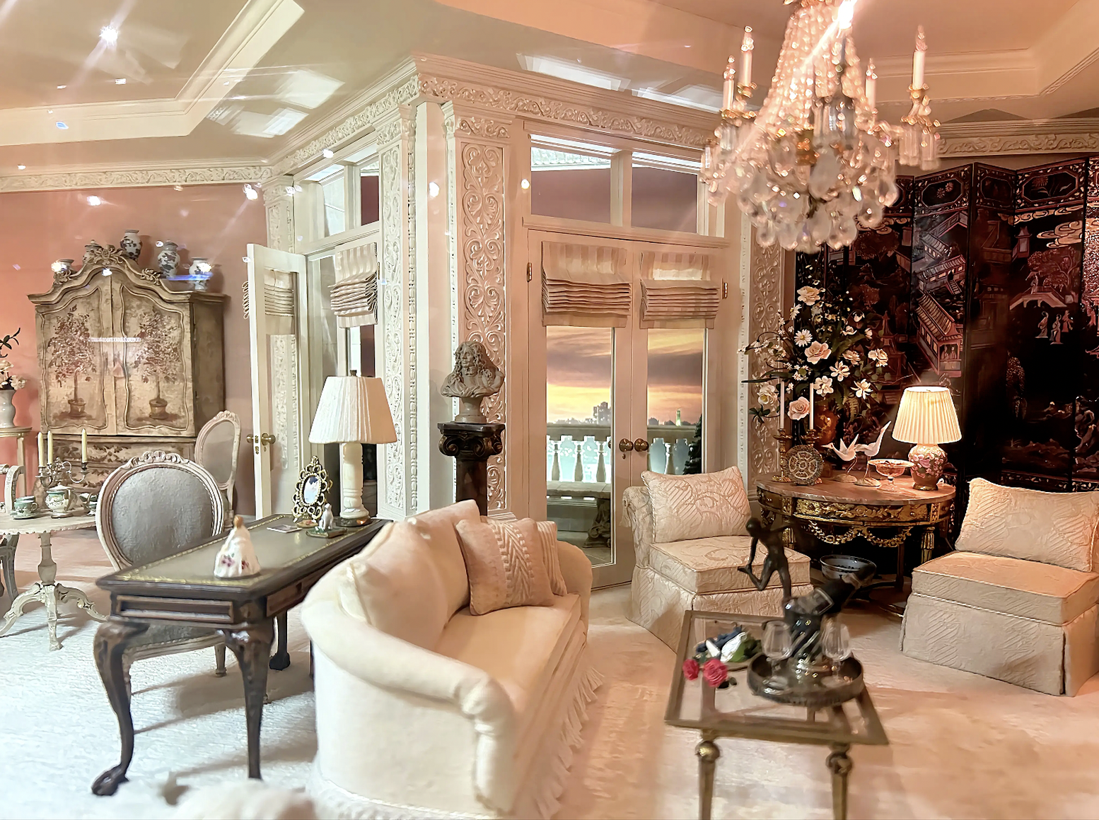 Luxurious pastel living room with ornate white carvings, a crystal chandelier, cream sofas, and antique furniture. Doors open to a balcony with sunset sky, flanked by flowers and decorative screens.