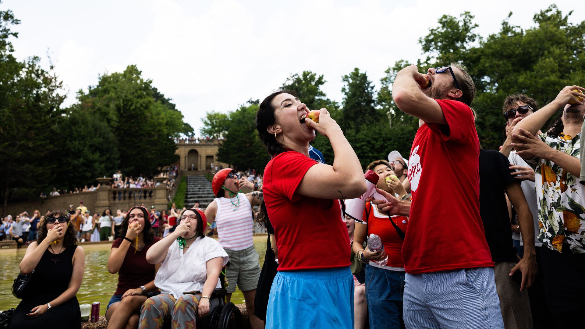 An outdoor crowd, many in red shirts, eats apples at a park. In the foreground, people bite apples while others watch. Green trees, blue sky, and a stone staircase with an arch in the background.