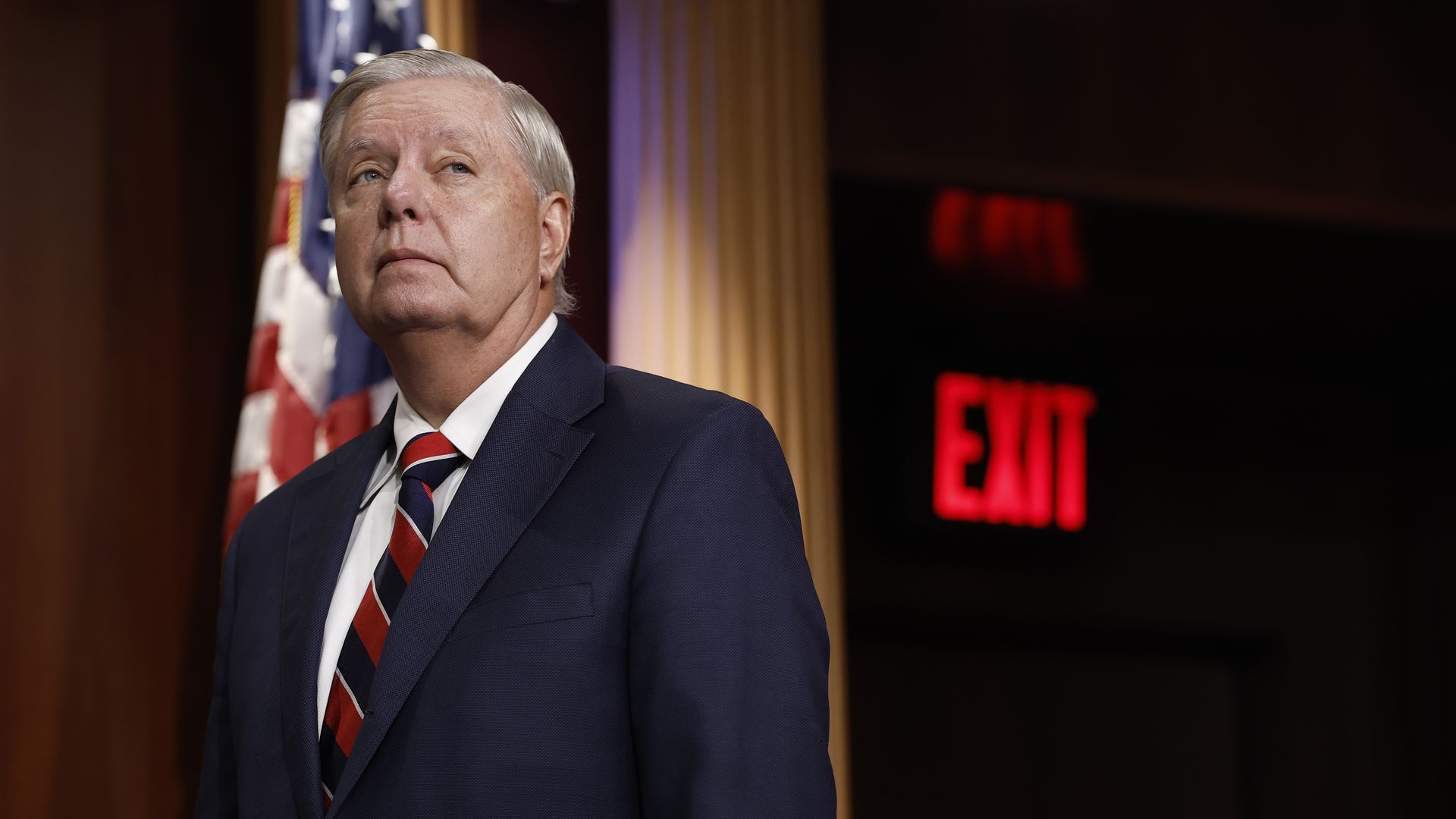 An older man in a navy suit and red striped tie stands on a stage, with an American flag behind him and a blurred red EXIT sign in the dark background.