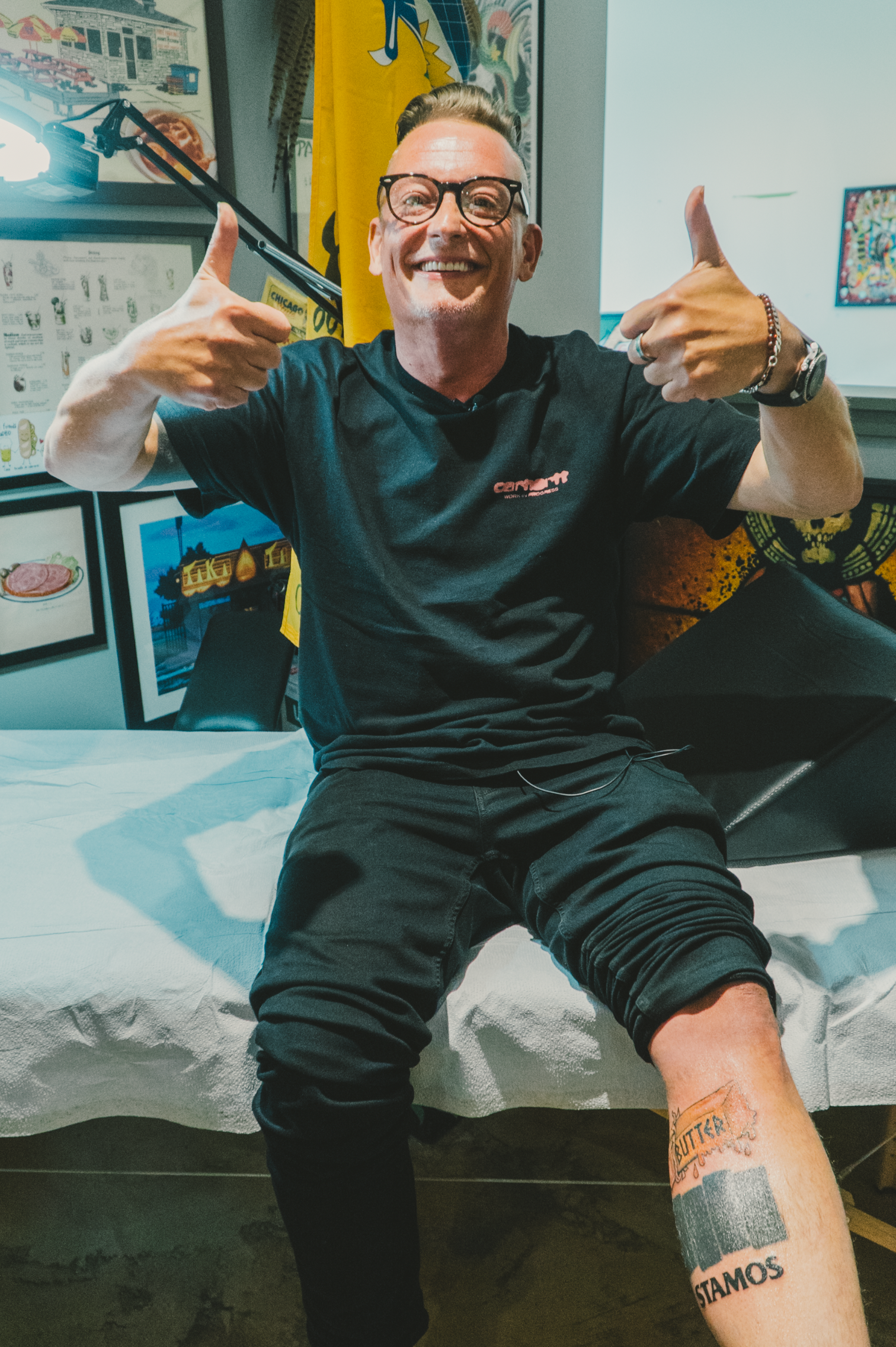 Man wearing black glasses and black clothes sits on a white-covered bench, smiling and giving thumbs up, showing tattoos on his left leg in a colorful, art-filled room.