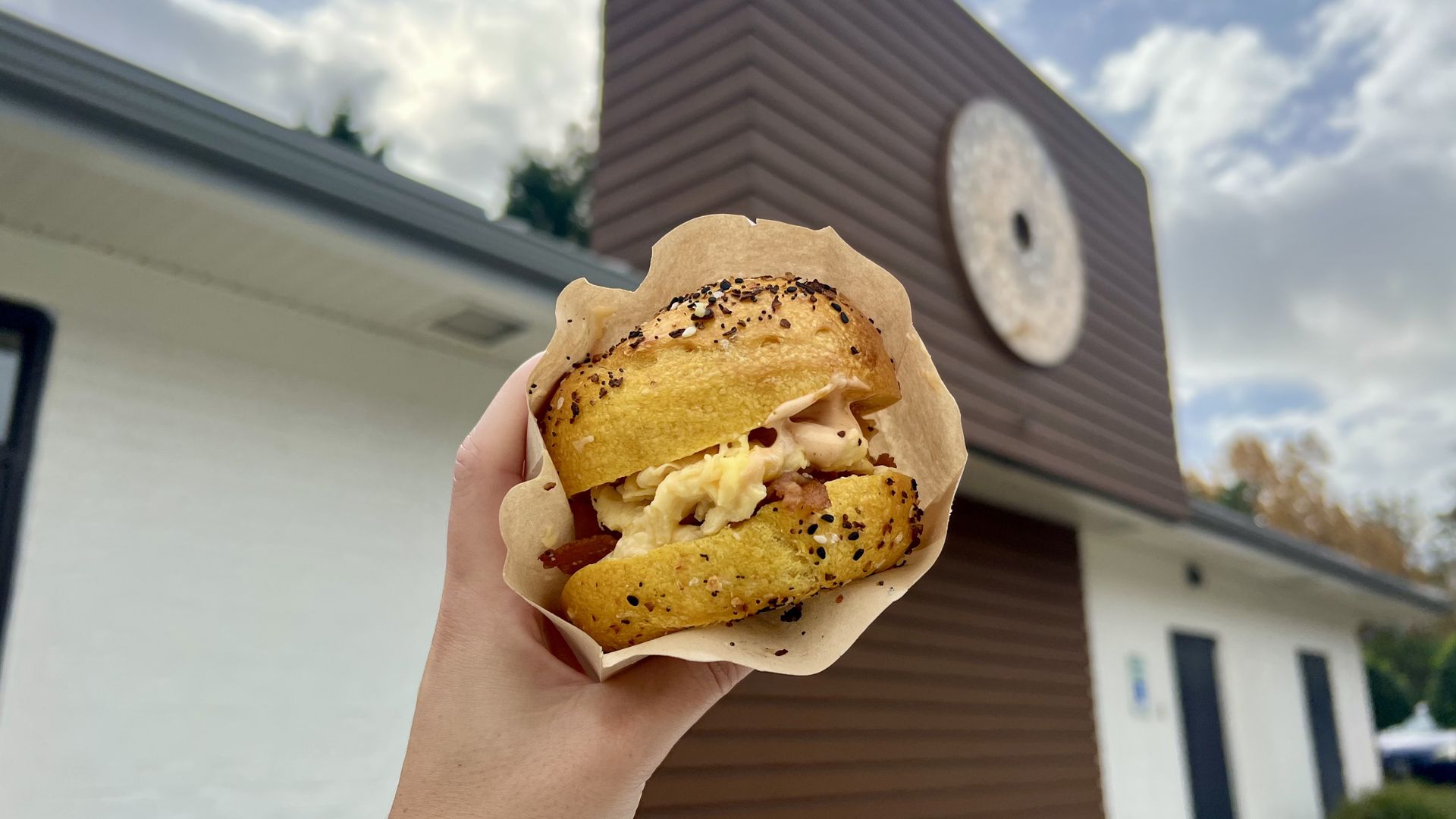 a hand holding up a everything bagel sandwich in a brown bag with egg poking out. In the background is a building with an everything bagel logo on it