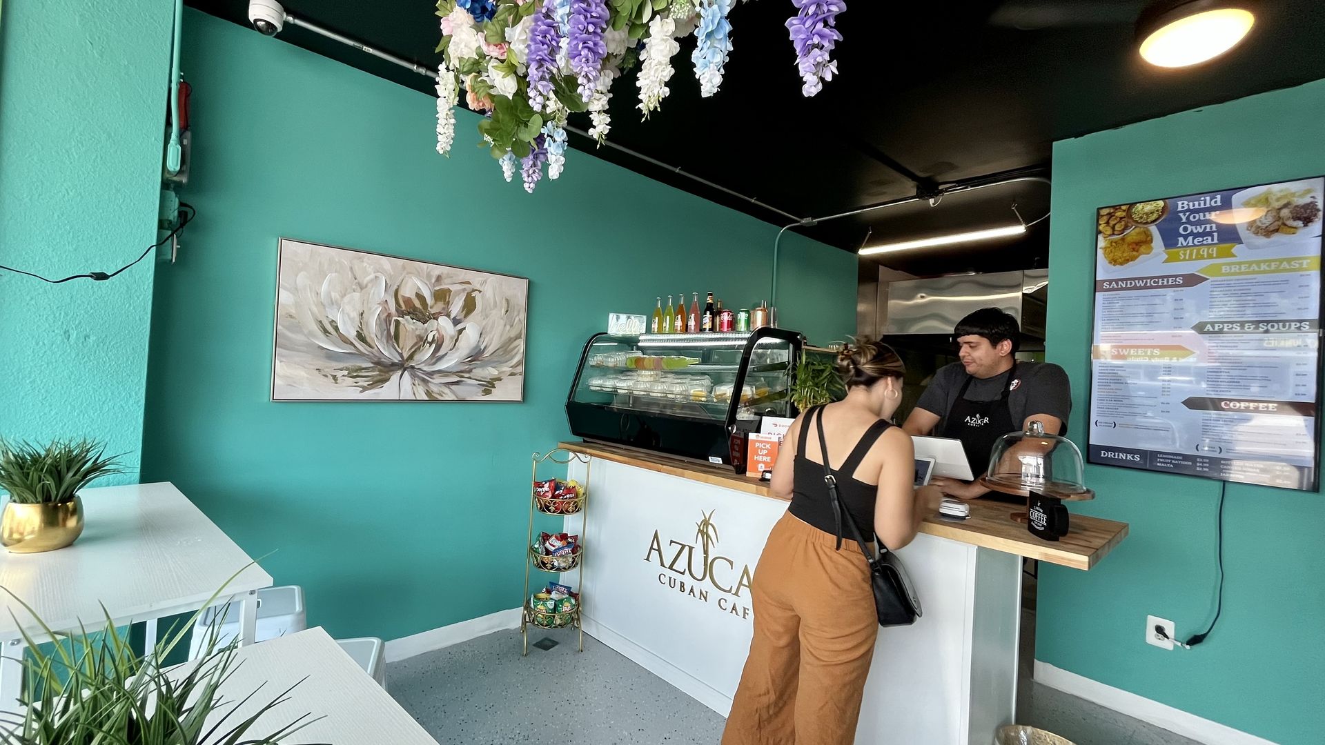 A man in a gray shirt and black apron takes the order of a woman in a black top and orange pants orders  at a white counter that says Azucar Cuban Cafe. The walls are bright teal, and purple, white and blue flowers hang from the ceiling.