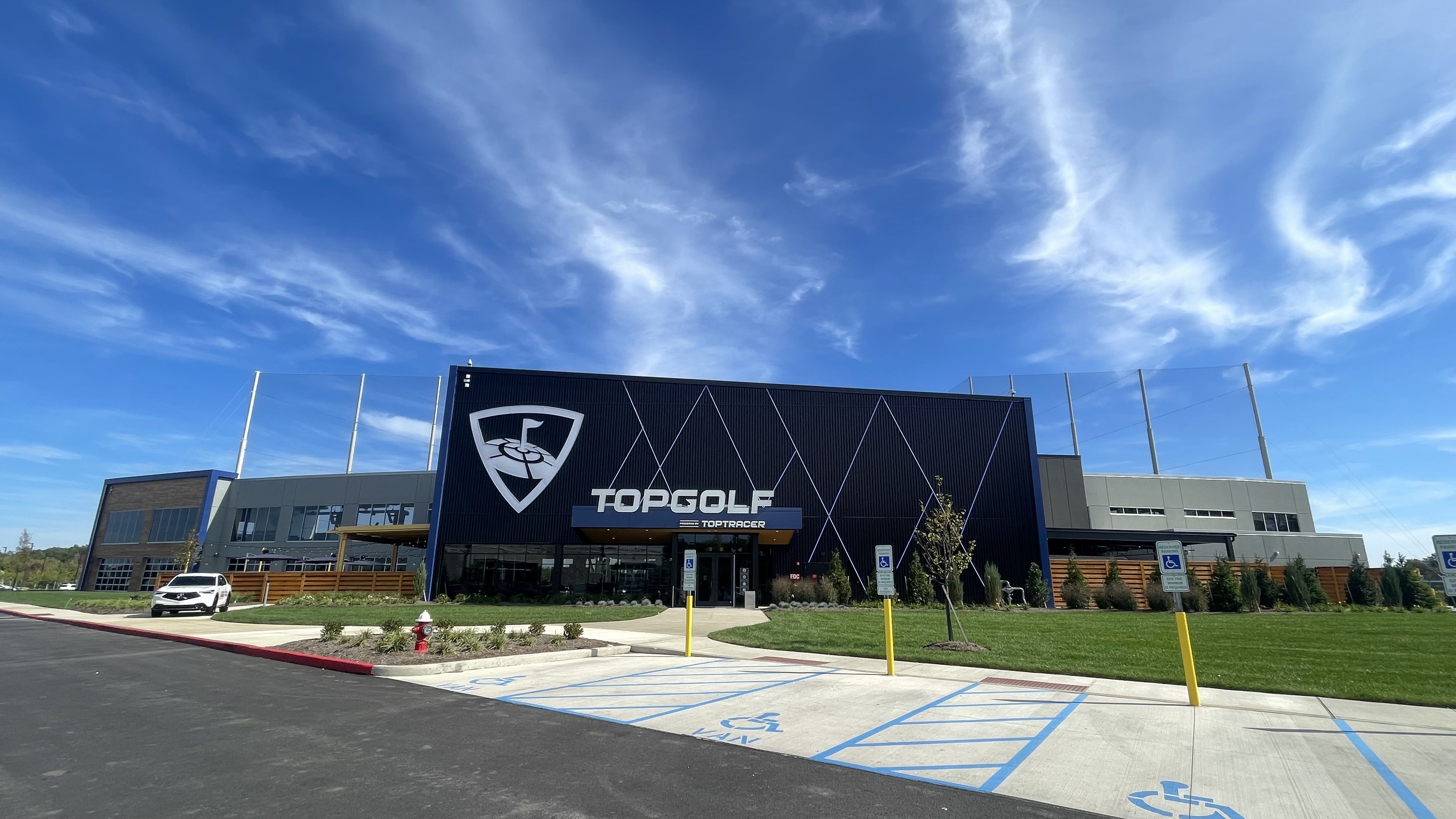 Front view of a modern Topgolf building under a bright blue sky with light clouds, adjacent to a handicapped parking area and green landscaping.