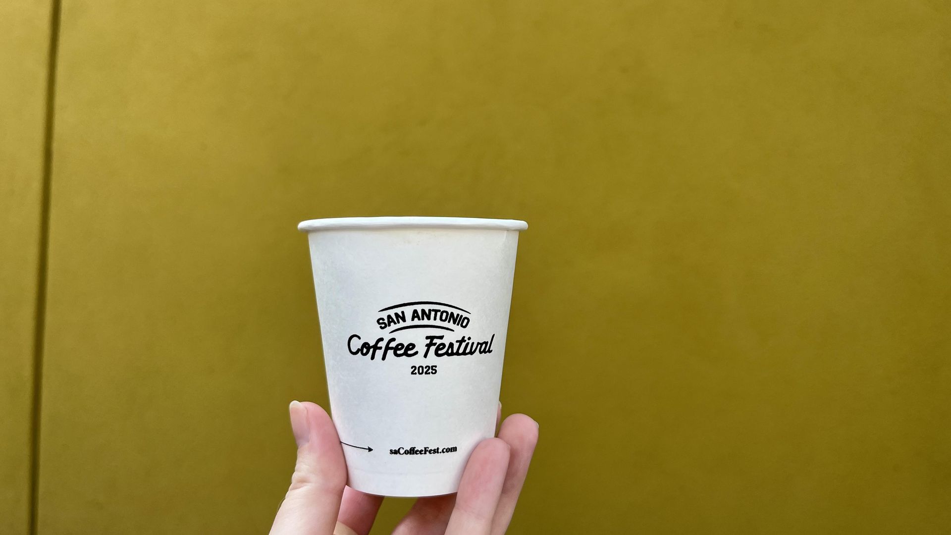 A small cup that says San Antonio Coffee Festival 2025, against a yellow wall back drop.