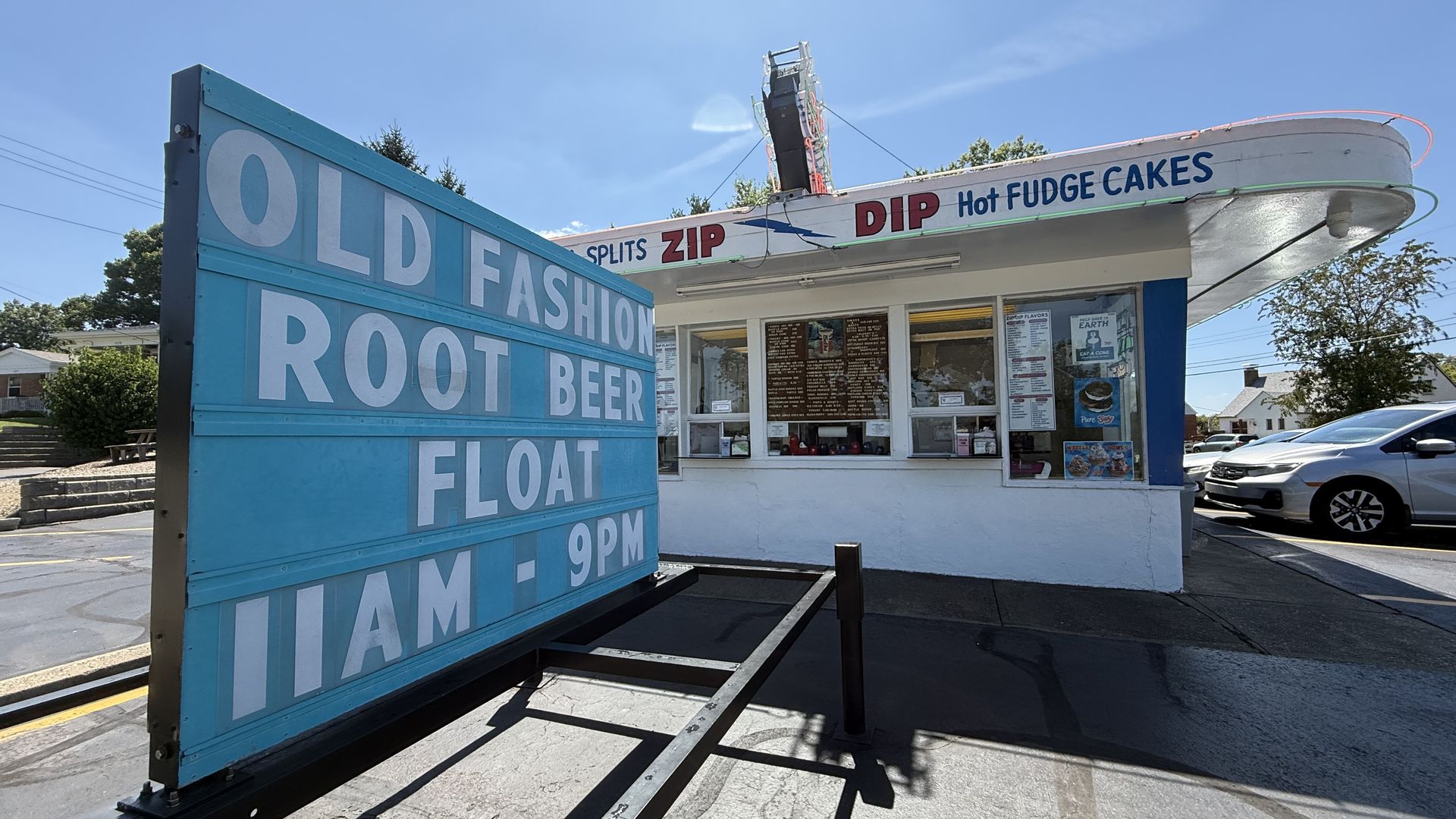 Bright blue roadside stand with a large white sign reading "OLD FASHION ROOT BEER FLOAT". Behind it, a white building advertises "DIP Hot FUDGE CAKES"; a car sits to the right under a clear blue sky.
