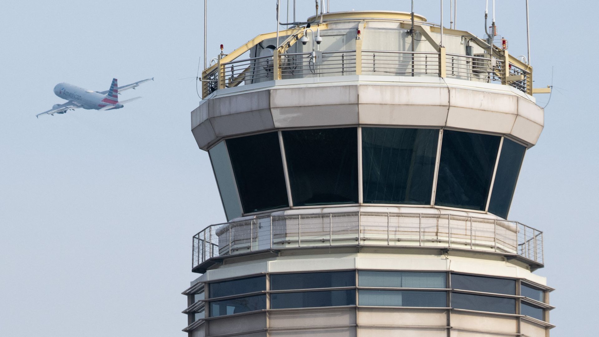 A close-up of a beige airport control tower with dark tinted windows and railings, and an American Airlines airplane flying in the pale blue sky on the left side of the image.