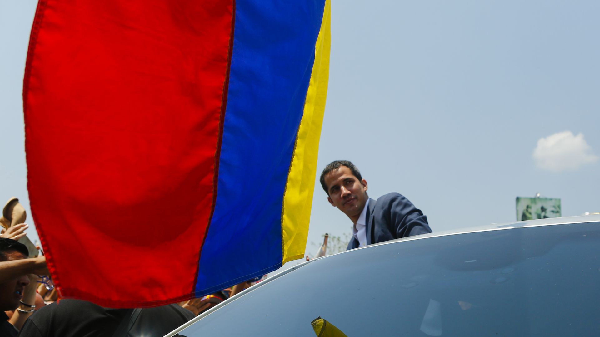 Juan Guaido stands next to a car with the Venezuelan flag hanging above him. 