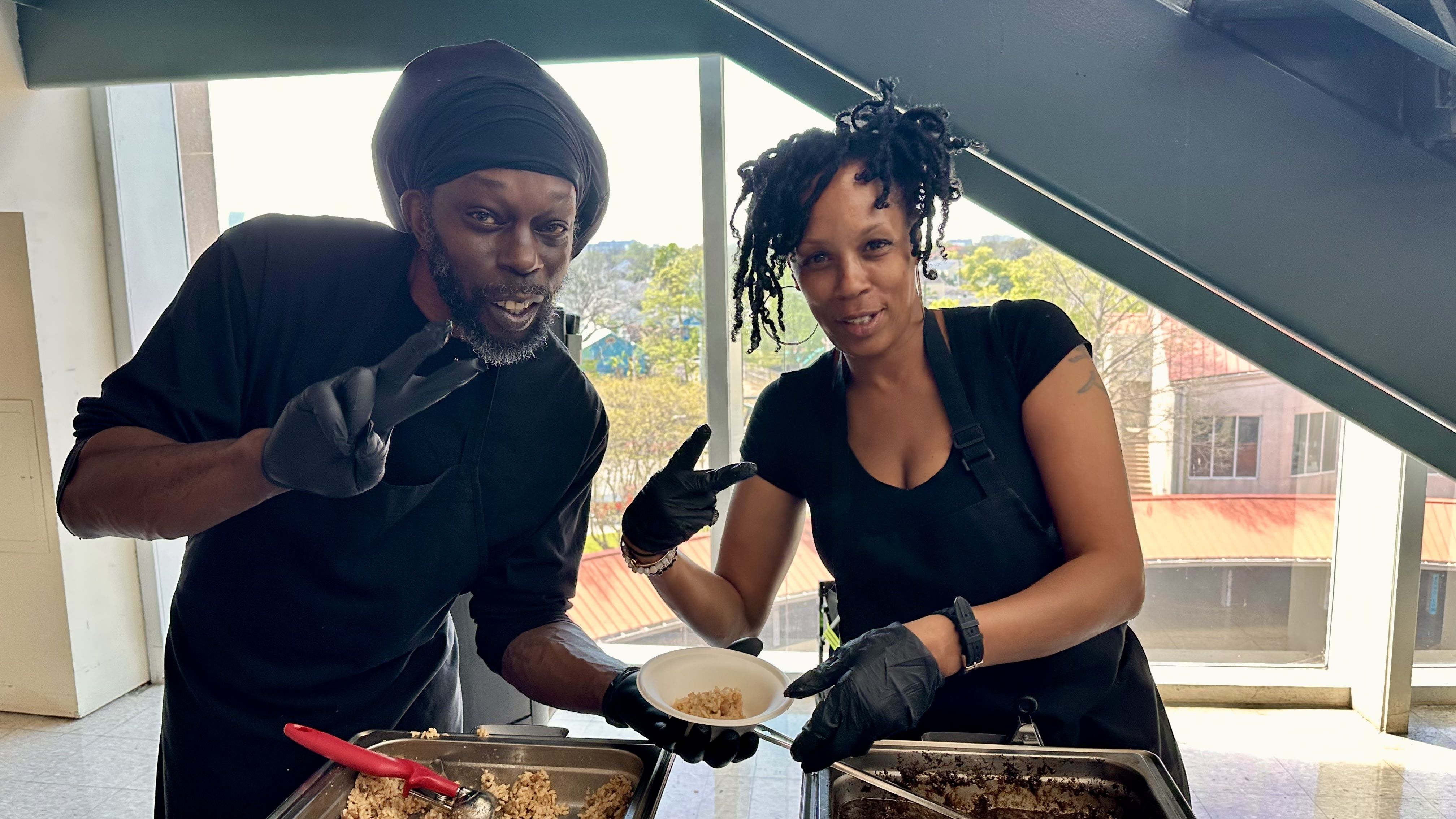 Two people in black uniforms and gloves pose with peace signs over metal food trays. Left: man wearing a black headwrap; right: woman with short dreadlocks. Bright kitchen with large windows behind.