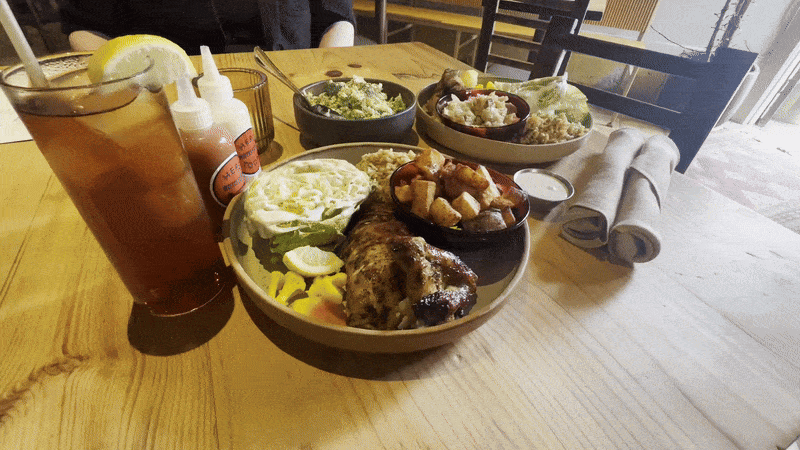 A camera pans toward two plates of rotisserie chicken with salad, pickled vegetables and roasted potatoes.