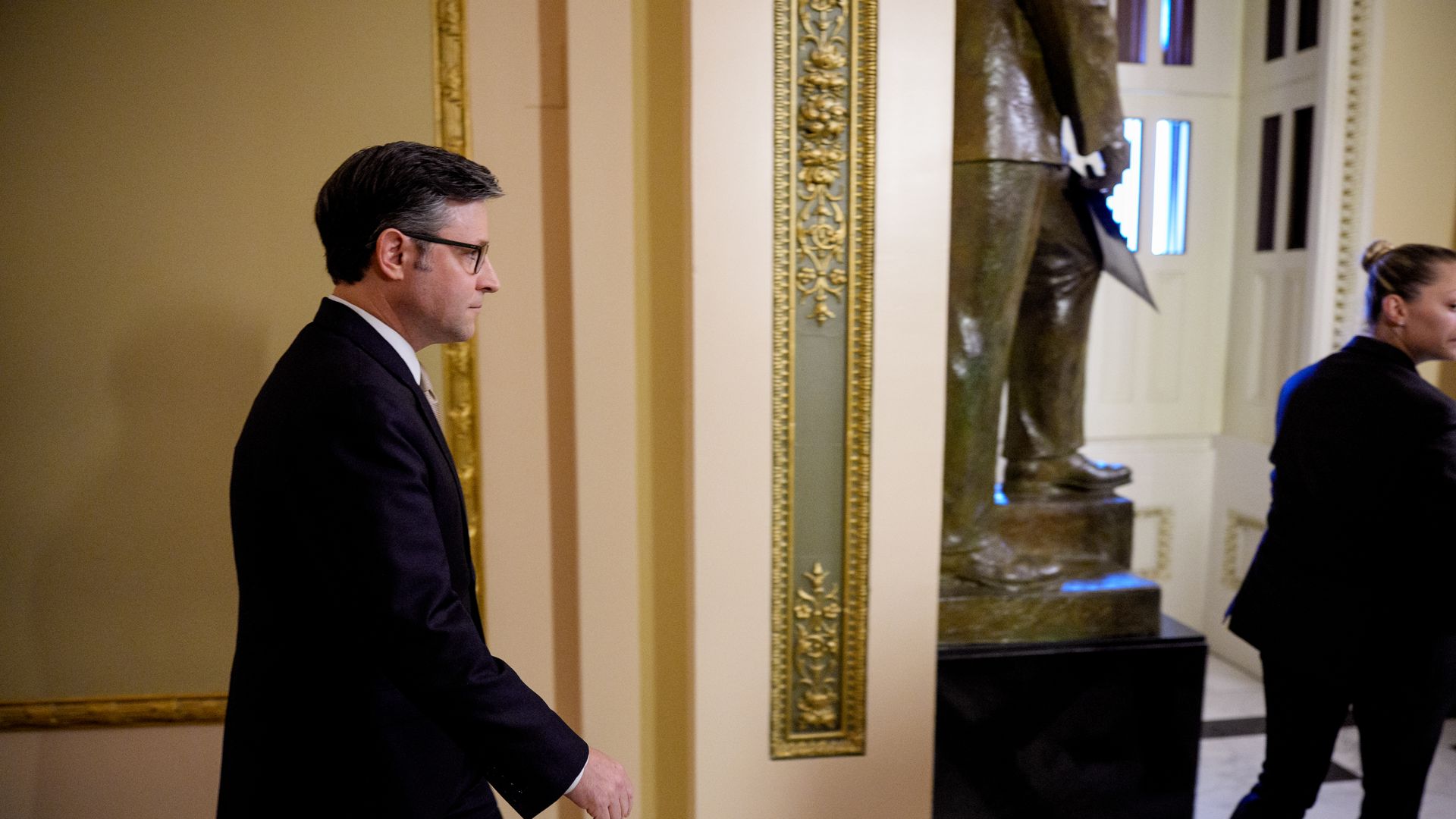 Man with glasses in a dark suit walking indoors past a large bronze statue and ornate gold-trimmed walls, accompanied by a woman in black attire walking ahead.