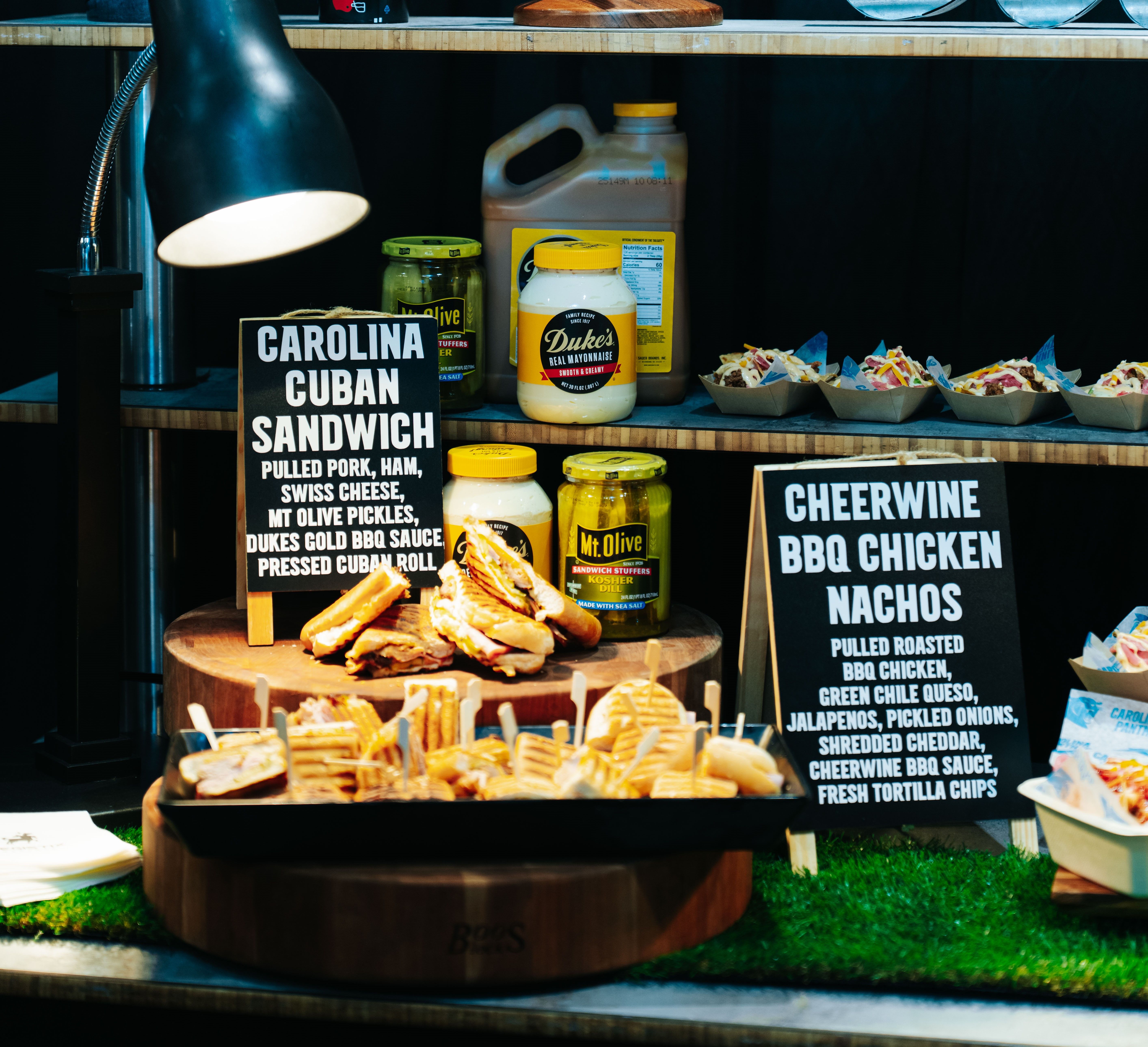Food display with Carolina Cuban Sandwiches and Cheerwine BBQ Chicken Nachos, jars of Mt. Olive pickles, Duke's mayonnaise, and BBQ sauce under a lamp on wooden shelves.
