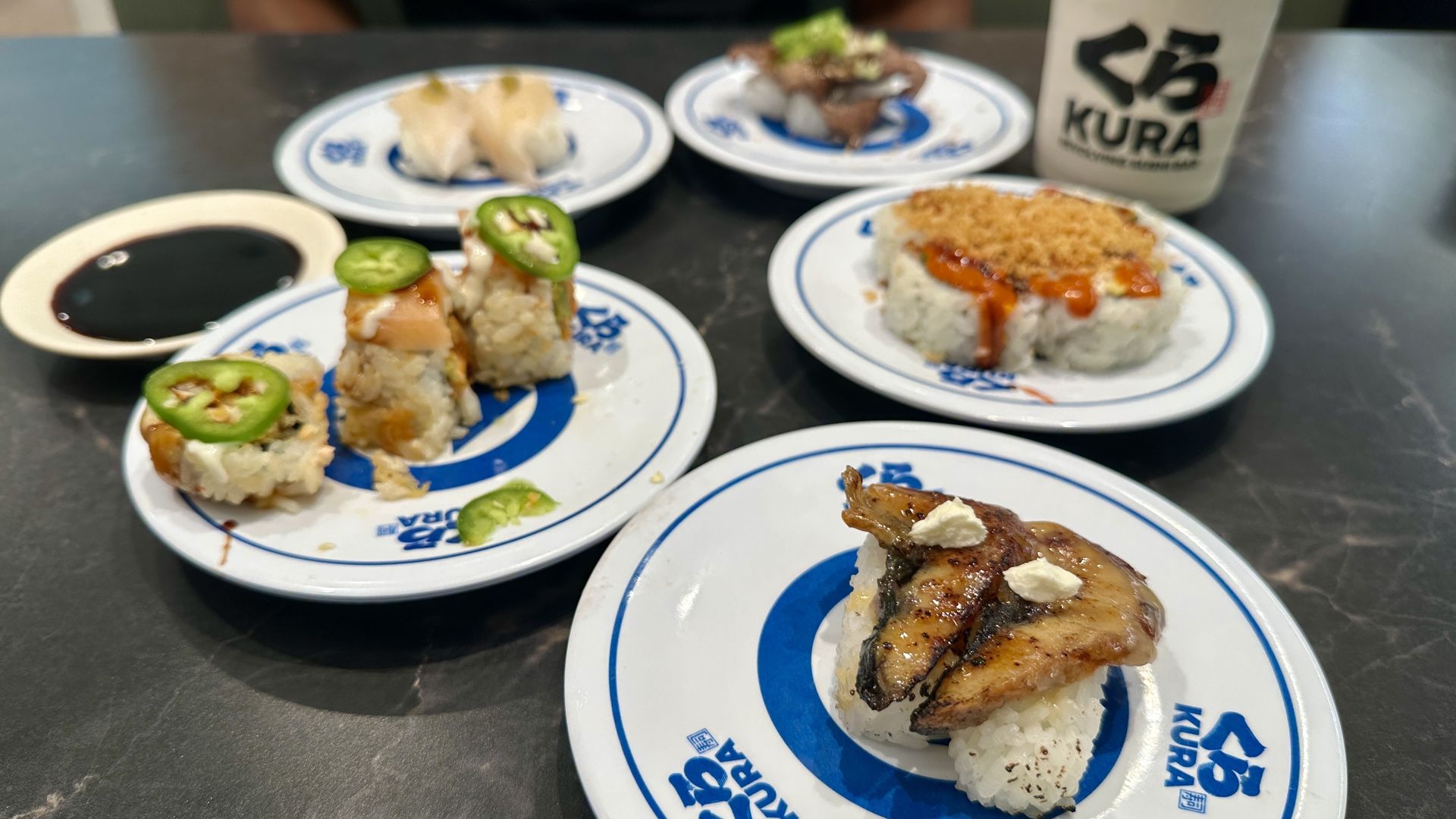 Assorted sushi plates on a dark table, topped with jalapeño slices and sauces; a white mug with the "KURA" logo sits in the background as a person is blurred behind the table.