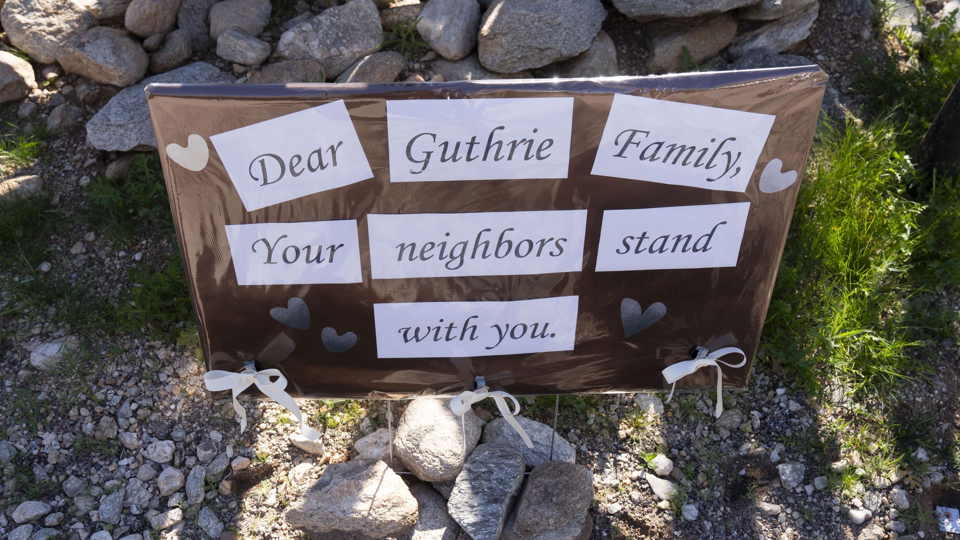 Brown sign outdoors among rocks and grass says, "Dear Guthrie Family, Your neighbors stand with you." with white bows and heart shapes.
