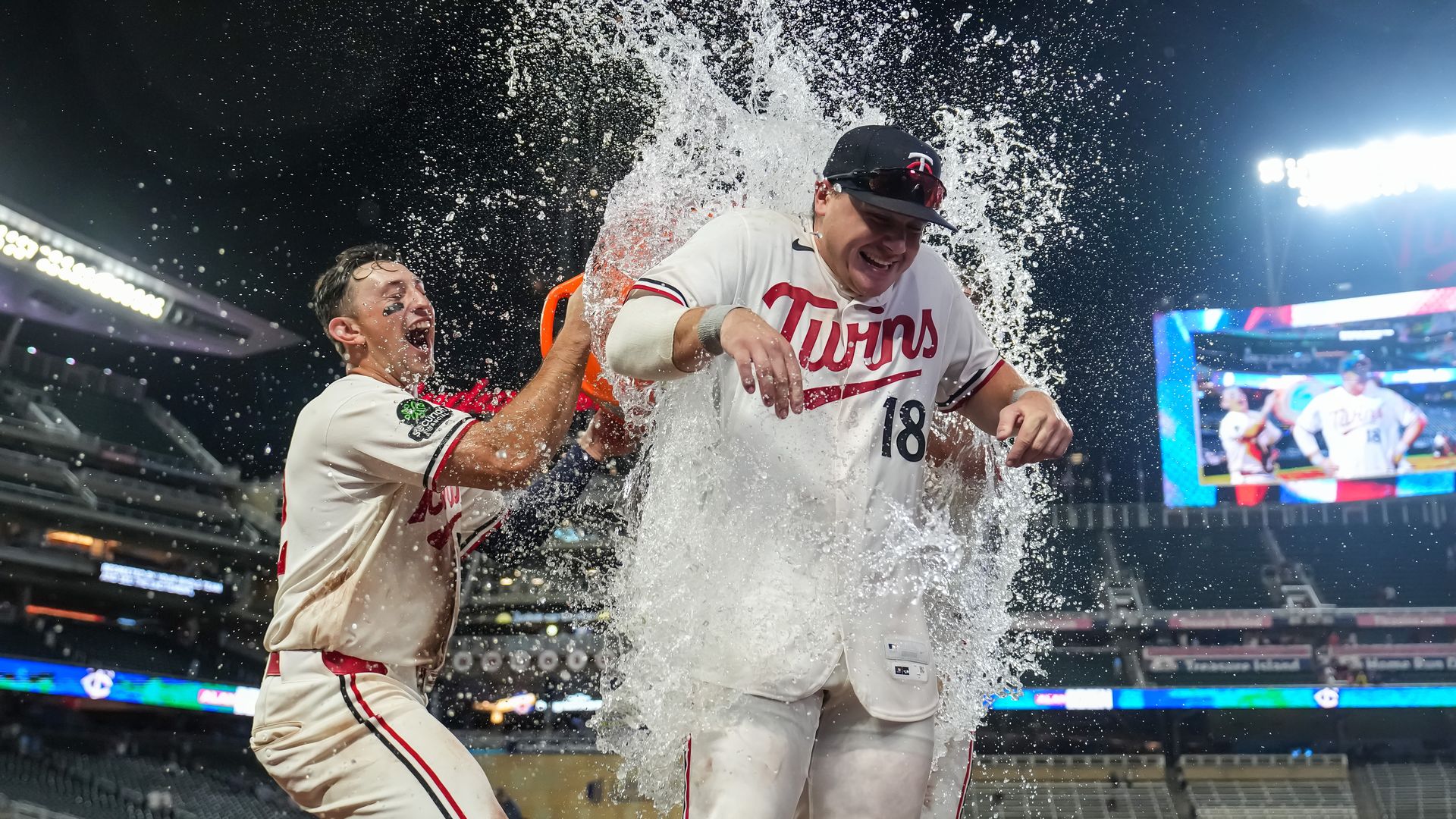 Two Twins players celebrate on the field as water erupts around them, wearing white uniforms with red trim; stadium lights and a large scoreboard glow in the background.
