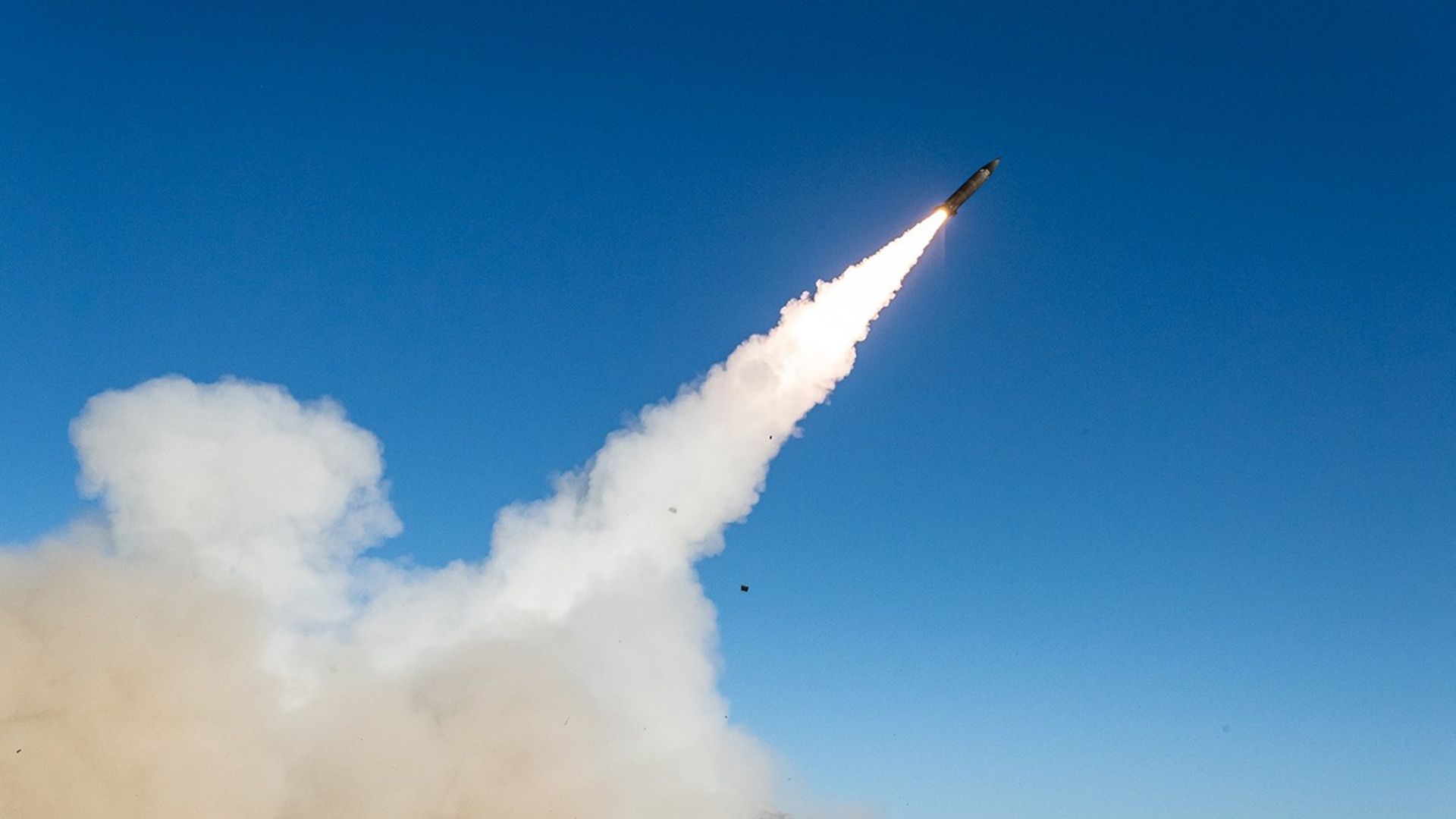 Missile launching into clear blue sky, leaving bright flame and thick white smoke; desert landscape with mountains in the background.