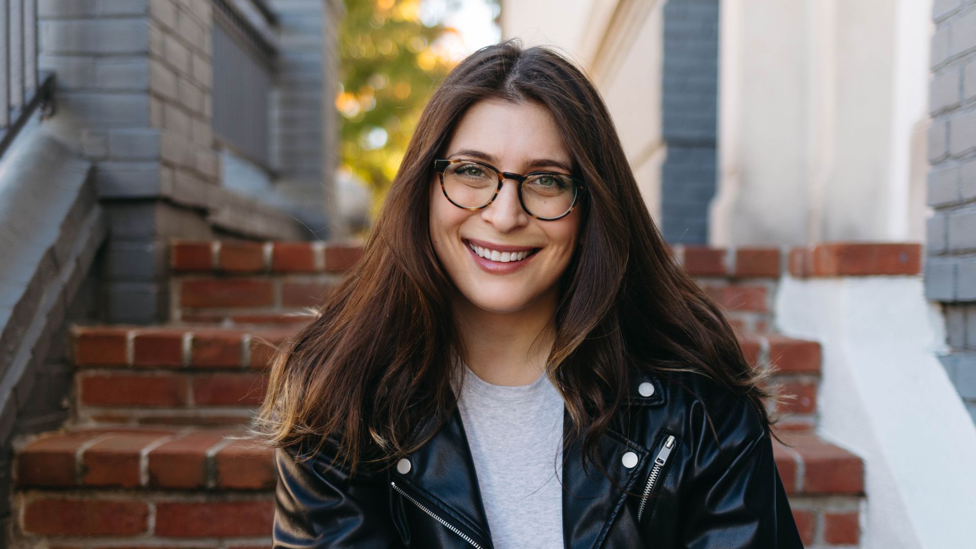 A photo showing a woman in a black leather jacket sitting on steps outside smiling.
