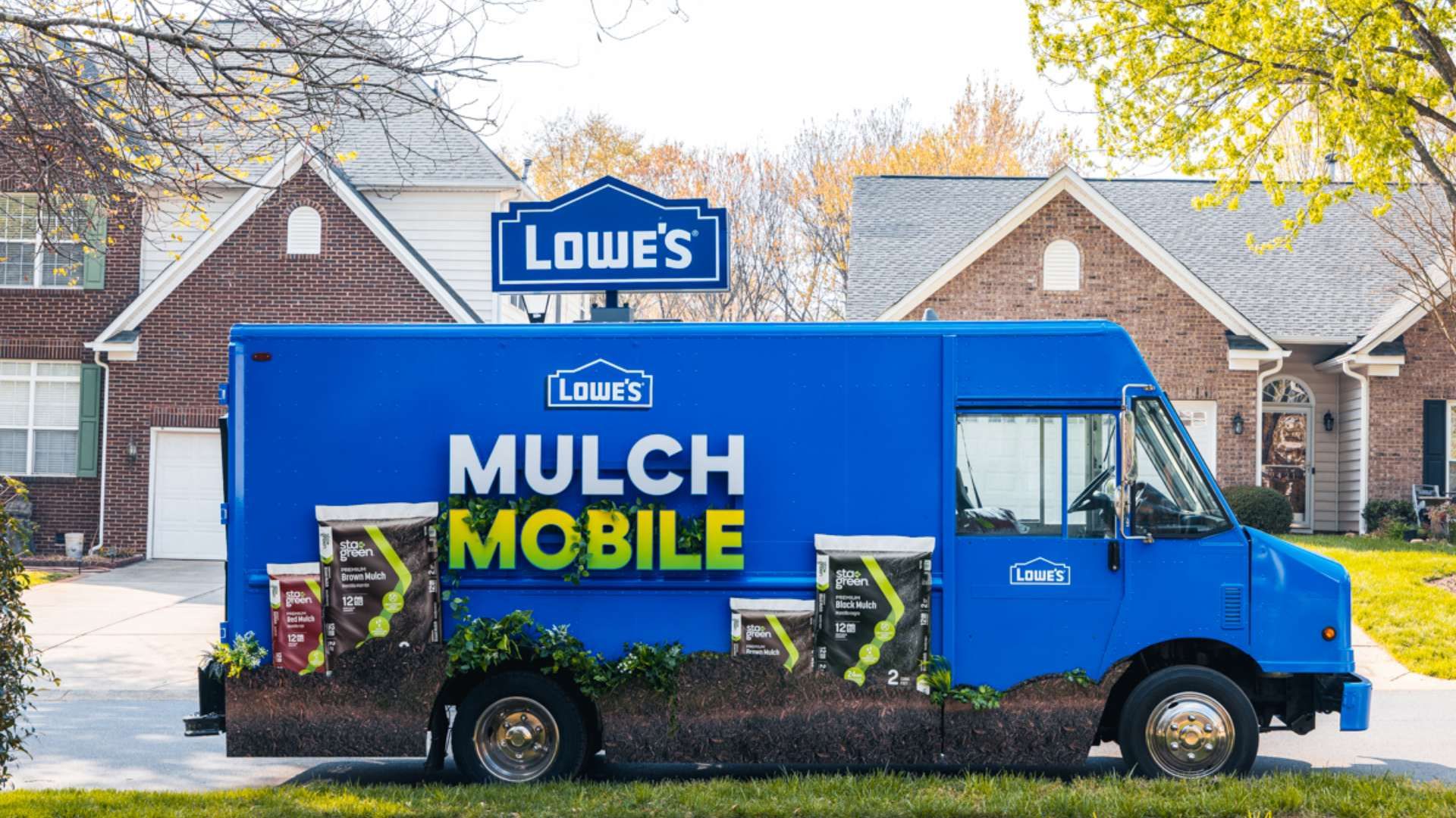 A bright blue Lowe's mulch delivery truck labeled "MULCH MOBILE" parked on a suburban street, with mulch bags at the side, a Lowe's sign, and brick houses in autumn trees.