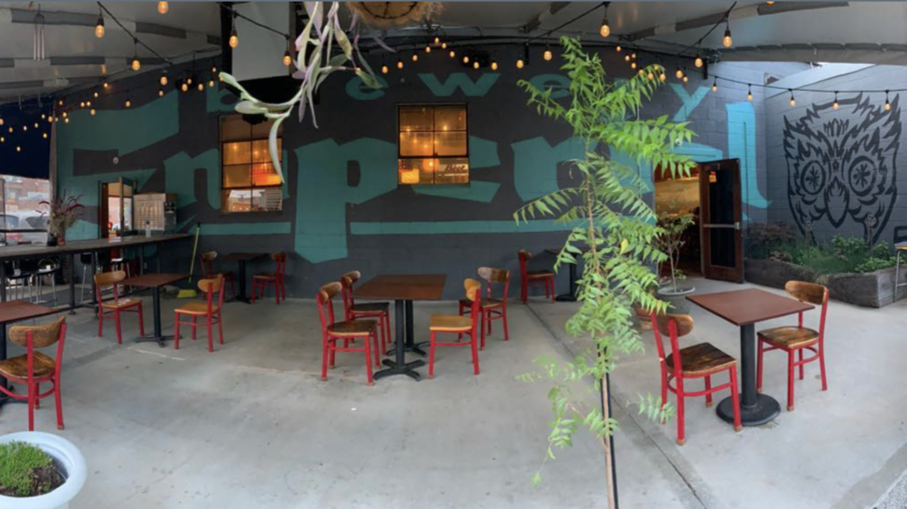 Open-air cafe with string lights, teal and gray mural walls, red metal chairs and wooden tables, potted plants, and a large owl graffiti mural on the right; warm orange light glows from the windows.
