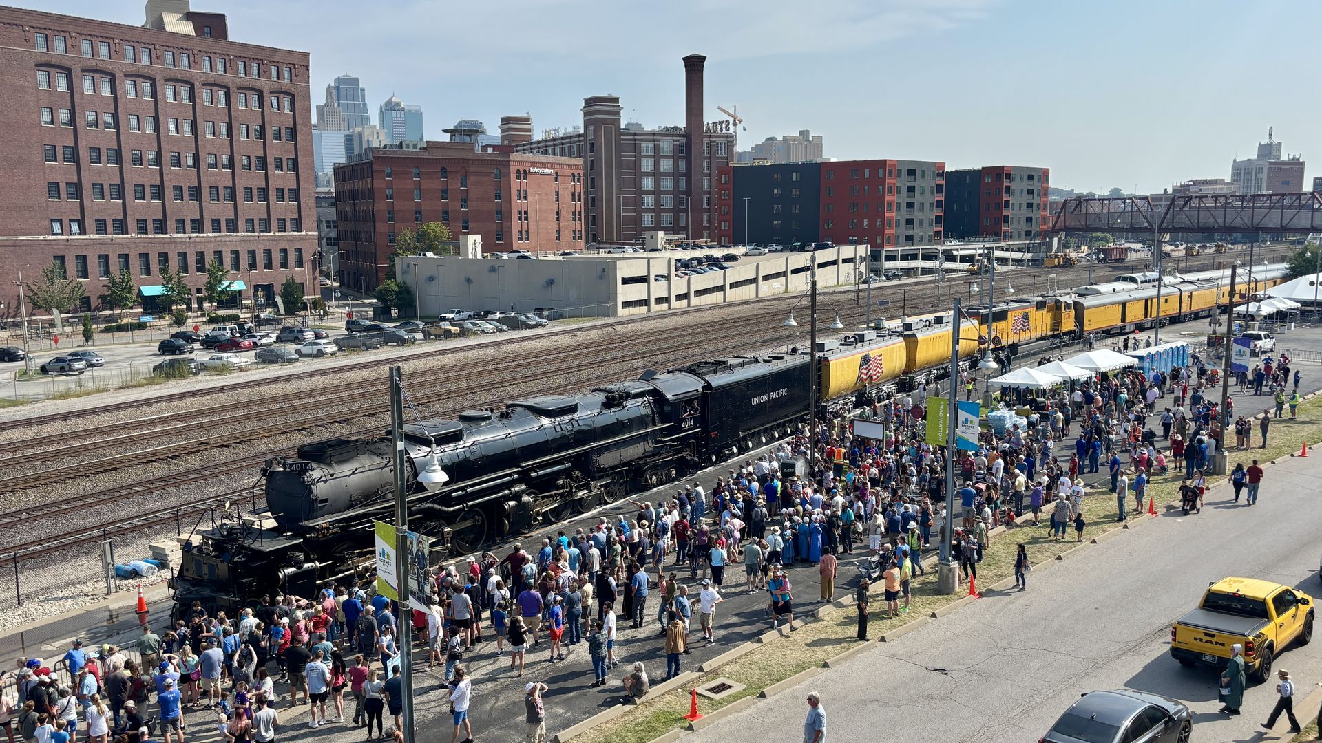 Large crowd gathers along a railroad yard to view a Union Pacific steam locomotive No. 4014 pulling yellow cars, with brick buildings and a distant city skyline in the background.
