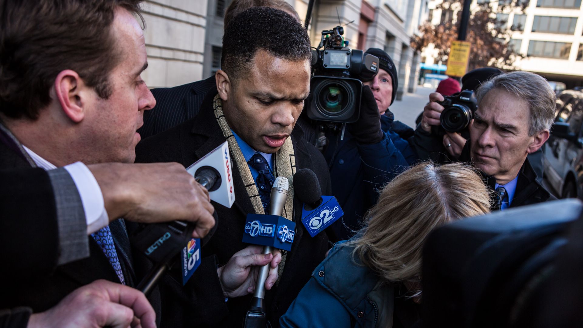 Photo of a man coming out of a building and talking to reporters who have surrounded him with microphones. 
