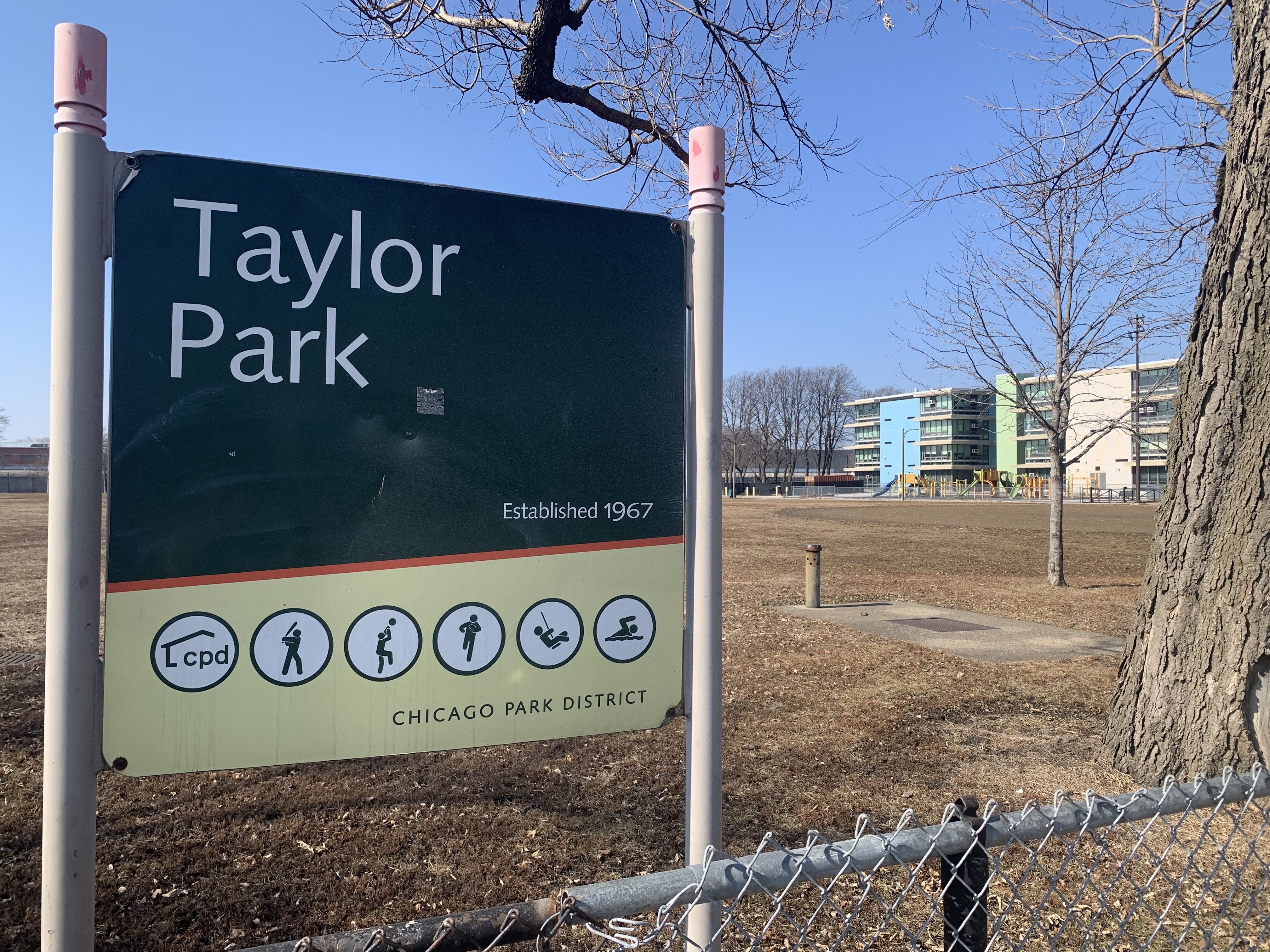 Green Taylor Park sign with white text and icons for activities, established 1967, in a dry grass area with leafless trees and a multi-story building under clear blue sky.