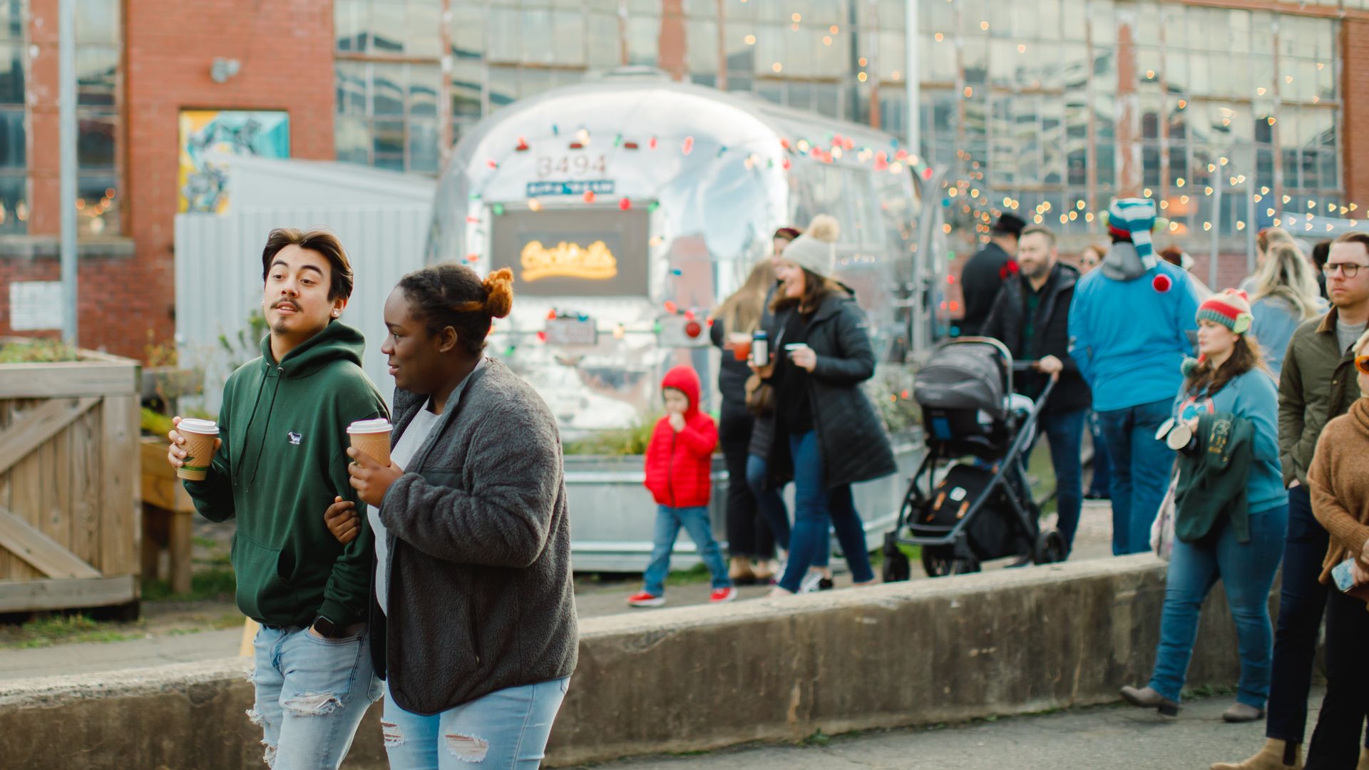 People wearing jackets and hats walk near a silver trailer decorated with colorful string lights outside an industrial building with large windows, holding warm drinks.