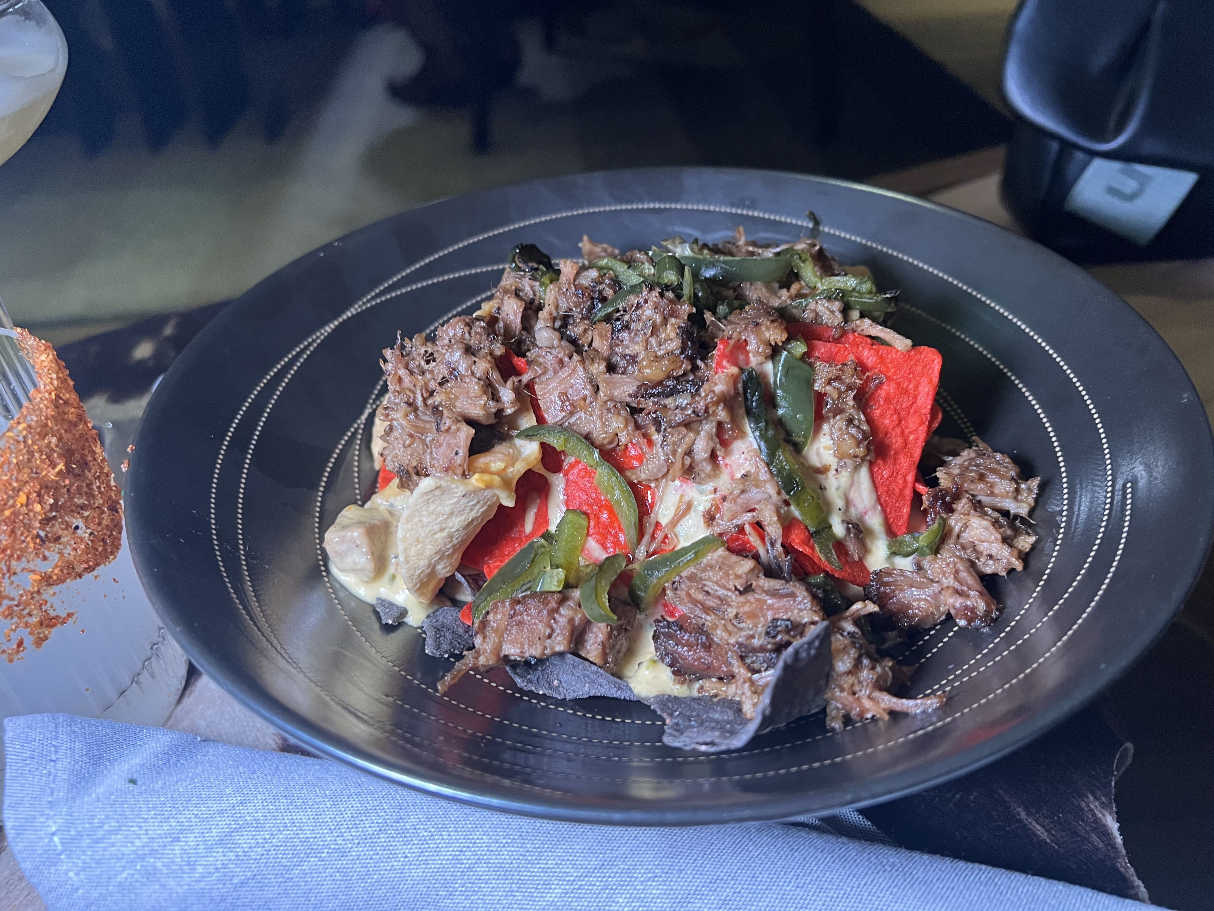 Black bowl of nachos with red and blue tortilla chips, shredded beef, green peppers, and melted cheese on a wooden table with a light blue napkin and a drink rimmed with chili powder.