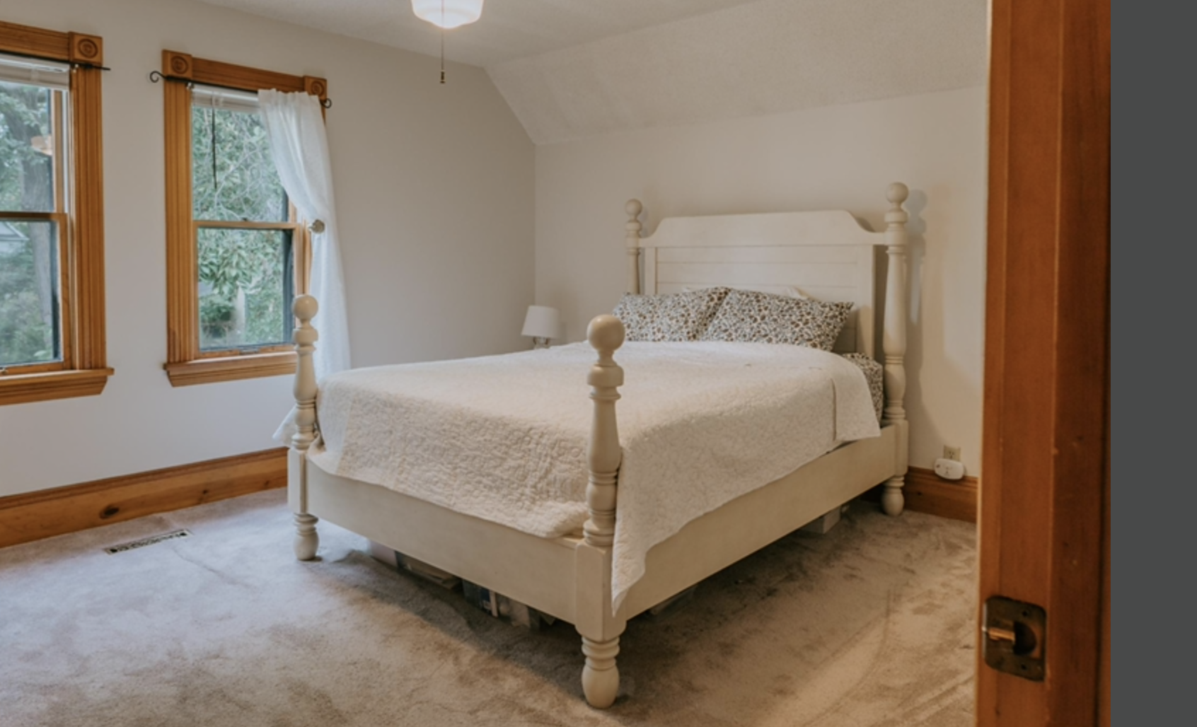 Cozy bedroom with a cream-colored wooden bed frame, white quilted bedding, patterned pillows, beige carpet, two windows with wooden trim, and a small side table with a white lamp.