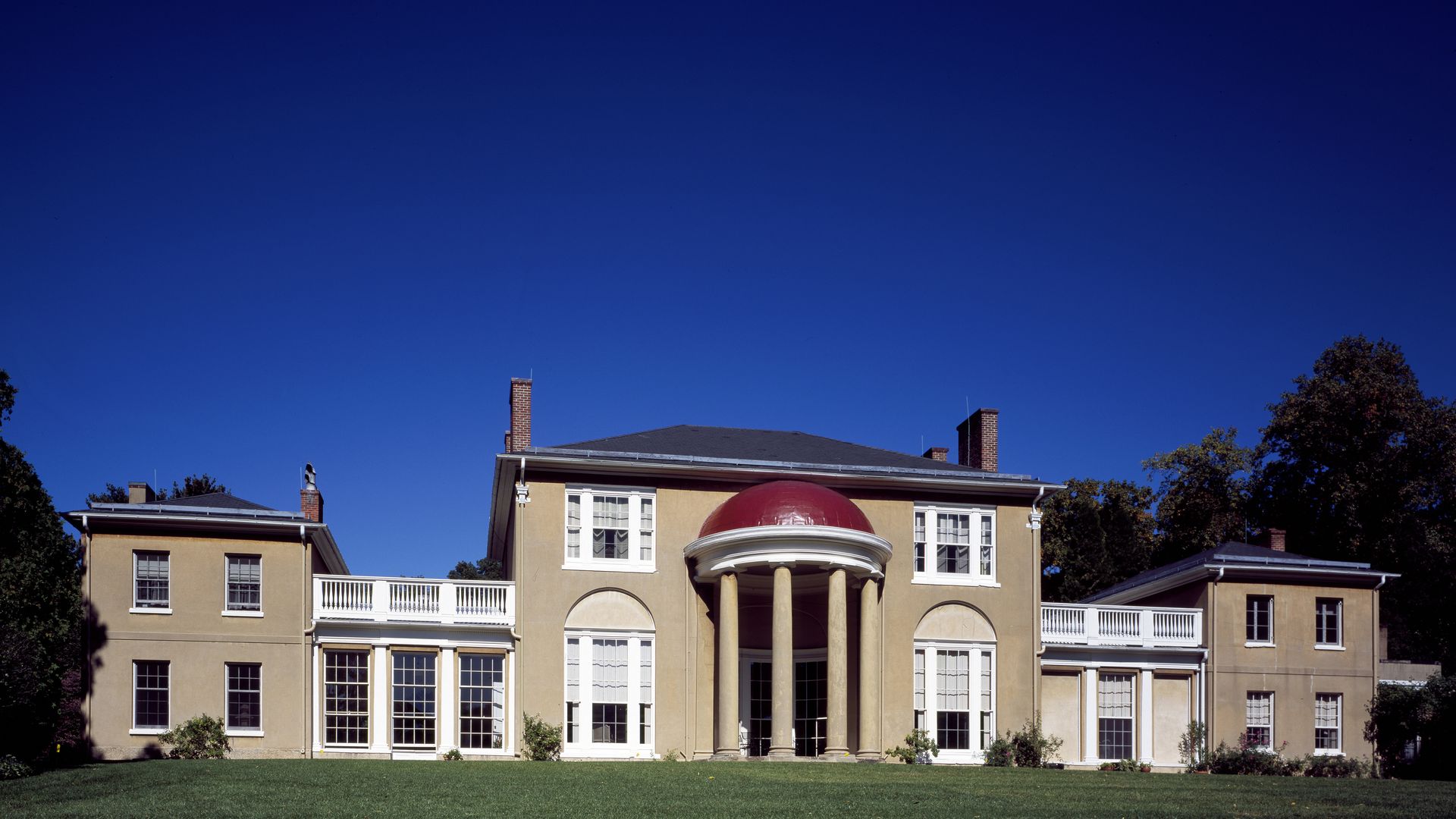 Beige, two‑story mansion with a central red domed portico supported by four columns, tall windows and white balconies. A bright blue sky and green lawn surround the building.