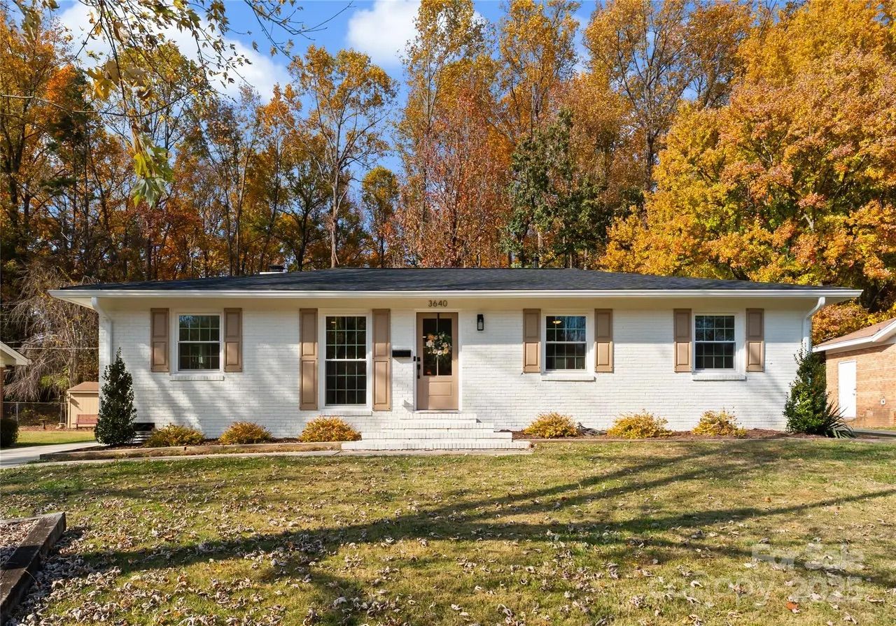 White brick single-story house with beige shutters and door, set against trees with autumn foliage in shades of yellow, orange, and brown, under a blue sky with clouds.