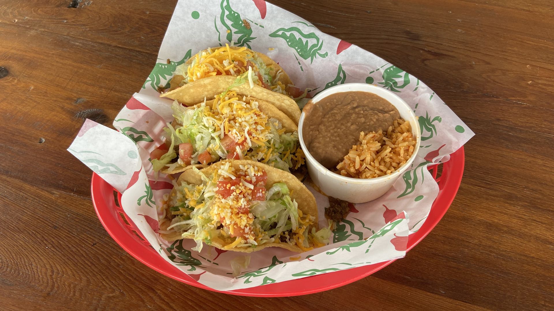Three cheese-covered tacos and a cup of refried beans in a red basket with white paper.