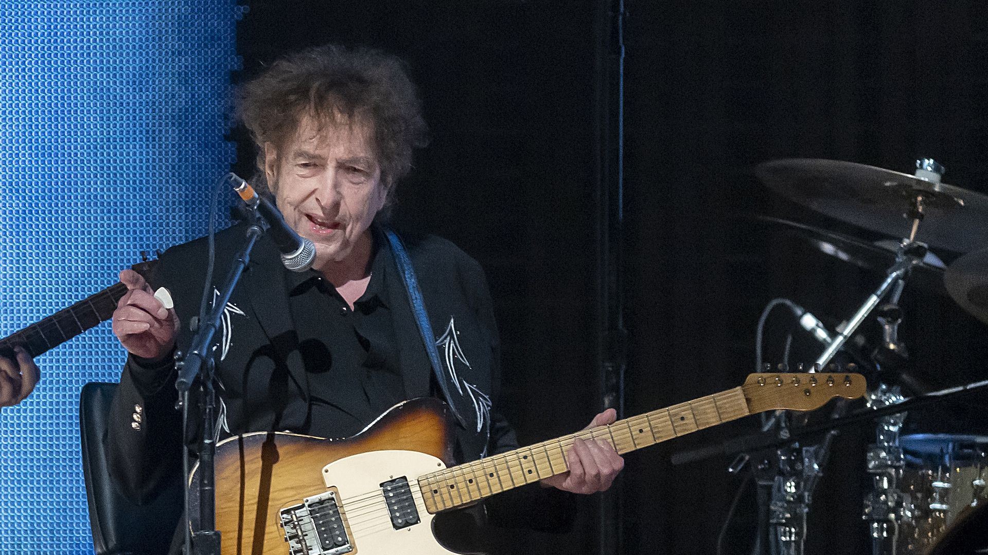 Bob Dylan performs on stage, sitting down with an electric guitar while pointing at the crowd. 