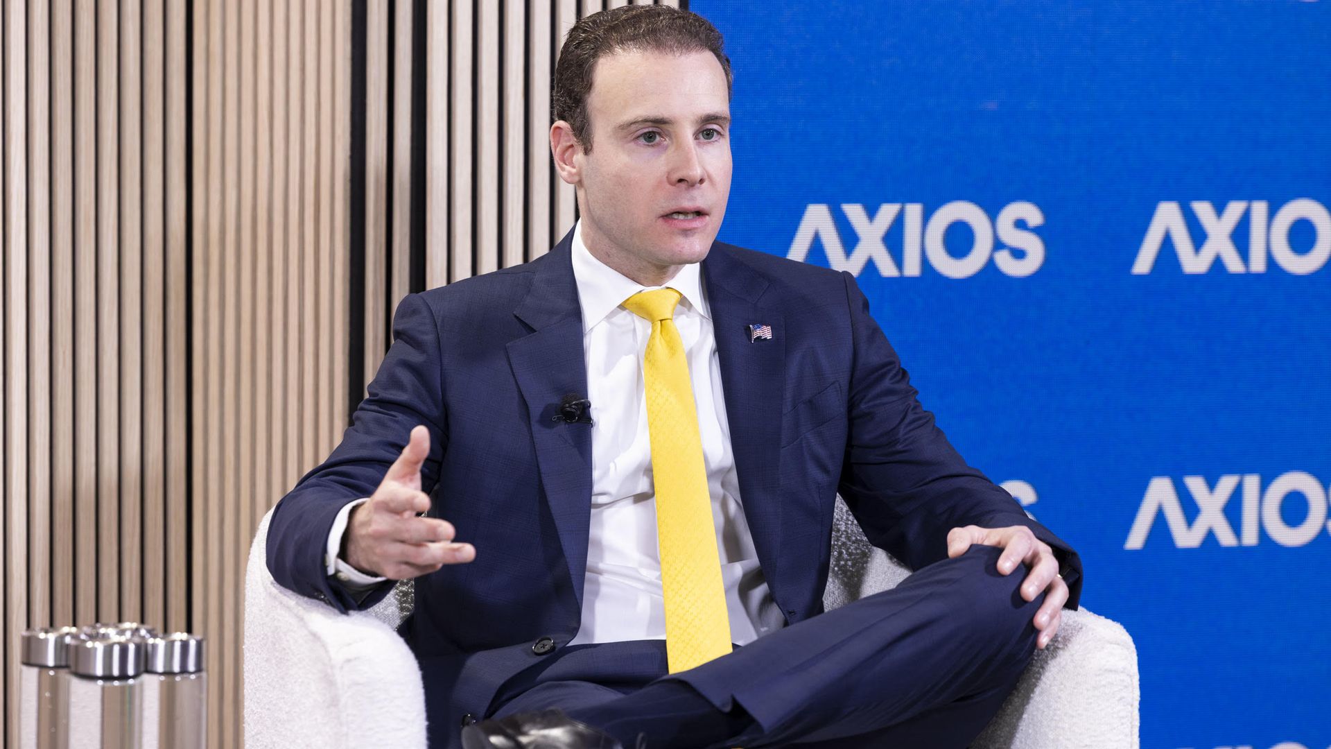A person in a navy suit with a yellow tie sits on a white chair, gesturing as he speaks. A blue Axios backdrop with white logos fills the background.