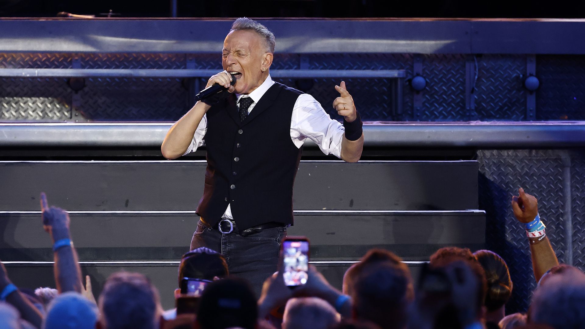 Male singer in white shirt and black vest performs on stage with a microphone; eyes closed, one hand pointing, audience phones raised in foreground, metallic stage backdrop behind.