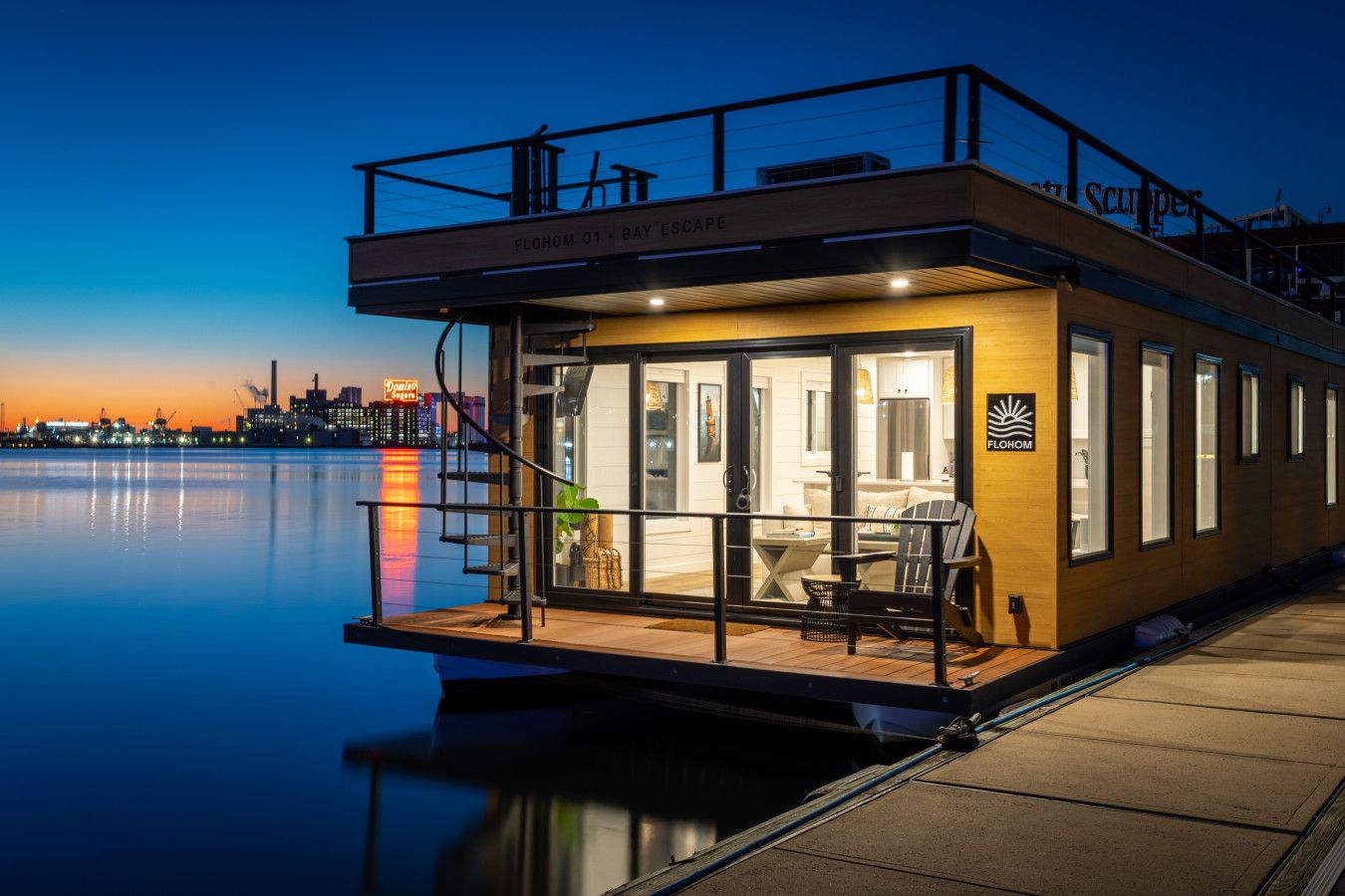 Floating houseboat with warm interior lights on a calm river at sunset; wooden exterior, glass doors, a small deck with seating, and distant city skyline glowing.