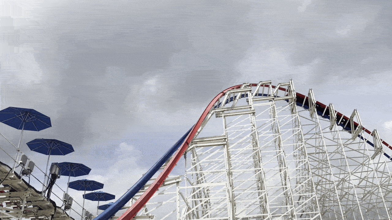 White lattice roller coaster with red and blue rails curves into a cloudy sky, while blue umbrellas shade a boardwalk on the left.