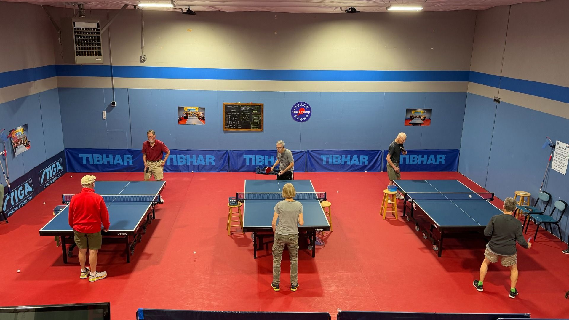 Indoor table tennis room with red floor and blue walls. Six older adults play doubles on three blue STIGA tables, surrounded by blue TIBHAR banners and wall signs, under bright lights.