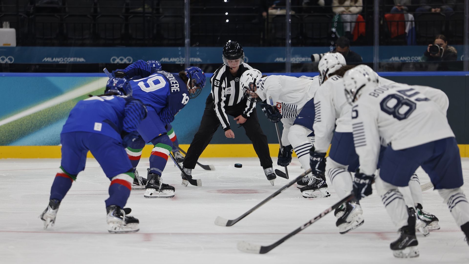 Ice hockey faceoff between players in blue and white jerseys with referee dropping puck on ice in an arena marked with Milano Cortina Olympic branding.
