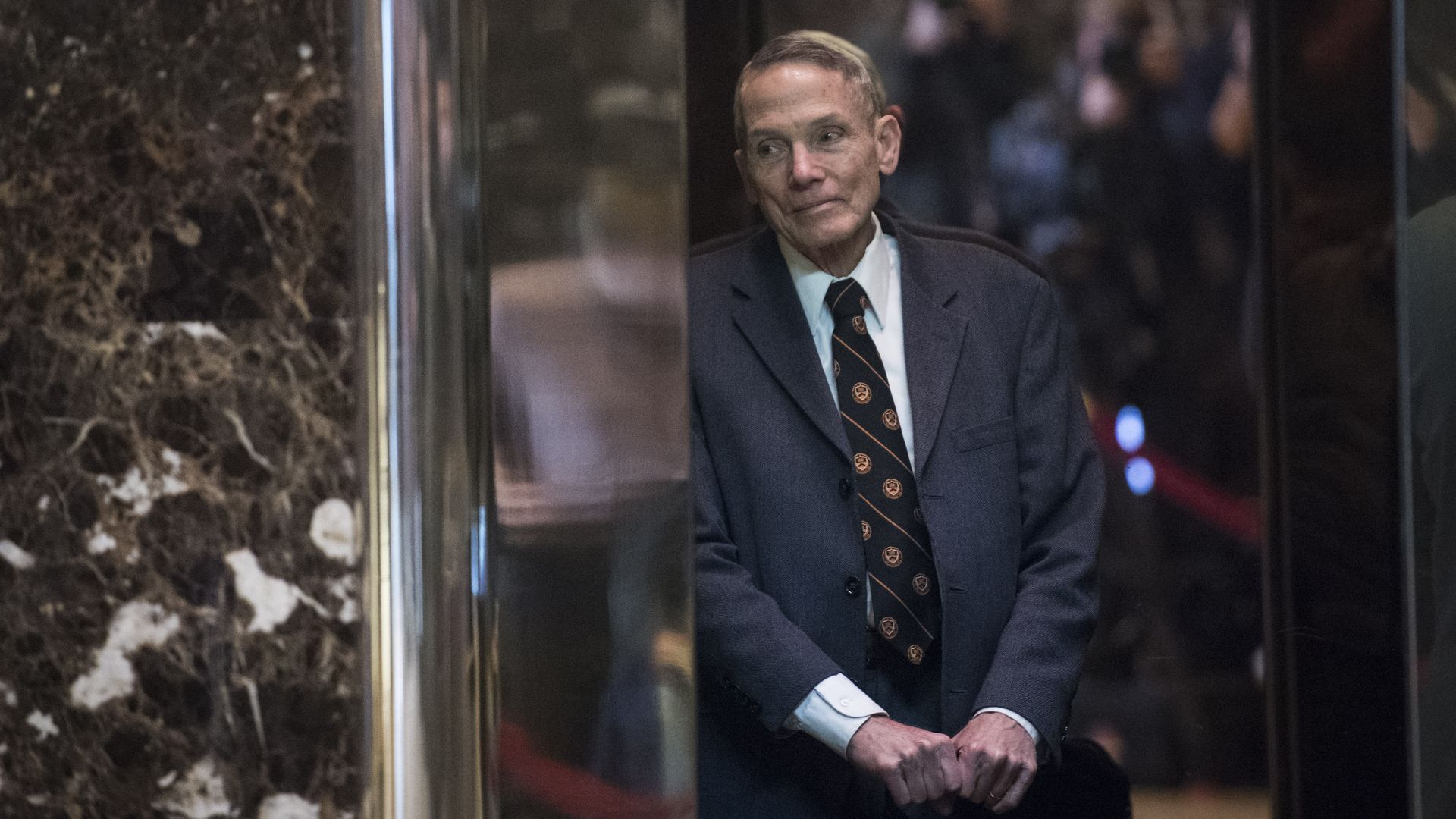 In this image, William Happer leans against a marble wall. He's wearing a gray suit and a black and orange striped tie.