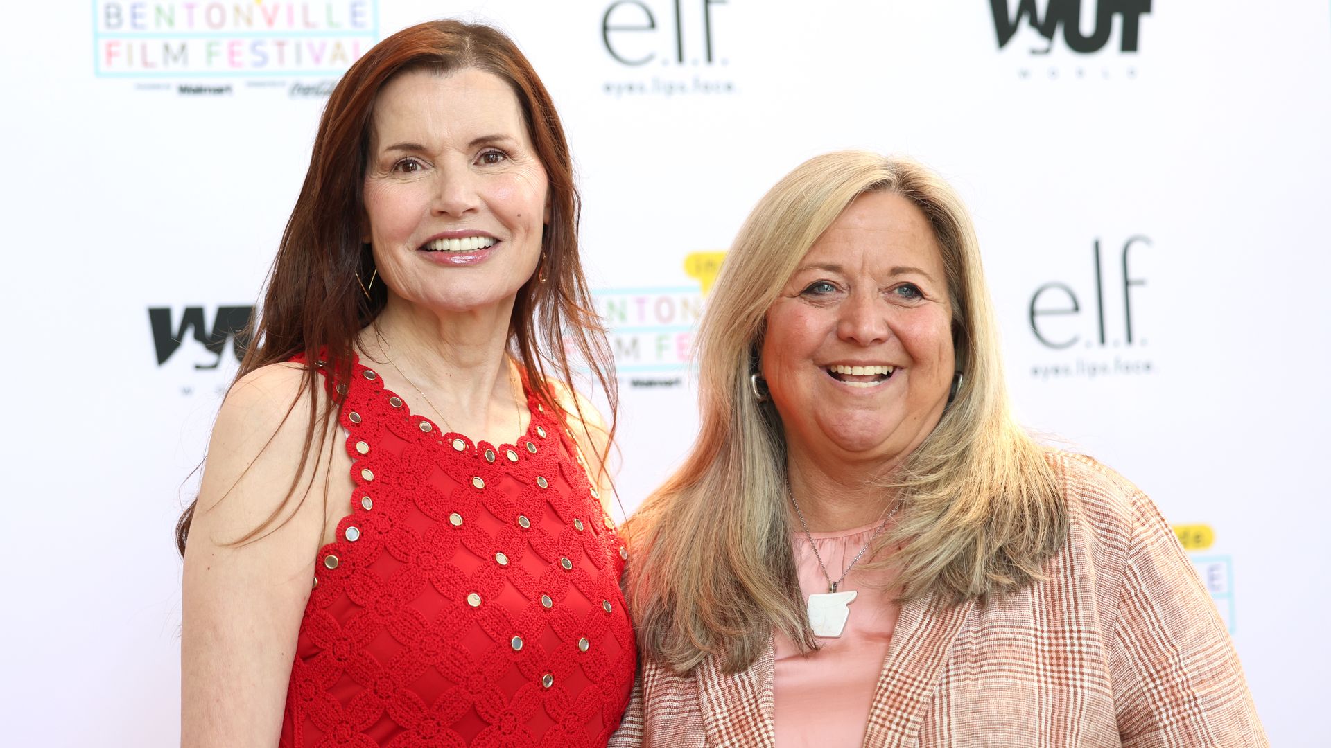 Two smiling women posing on a white backdrop at a film festival; left woman in bright red lace dress, right woman with blonde hair wearing a pink top and beige plaid jacket, logos visible behind.