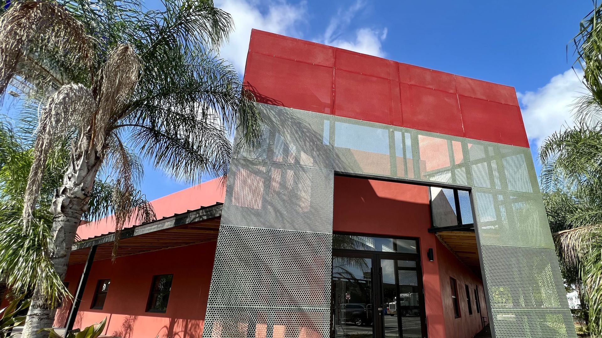 A modern red building with perforated metal panels and a bold red roof, glass entrance, and palm trees in the foreground against a bright blue sky.