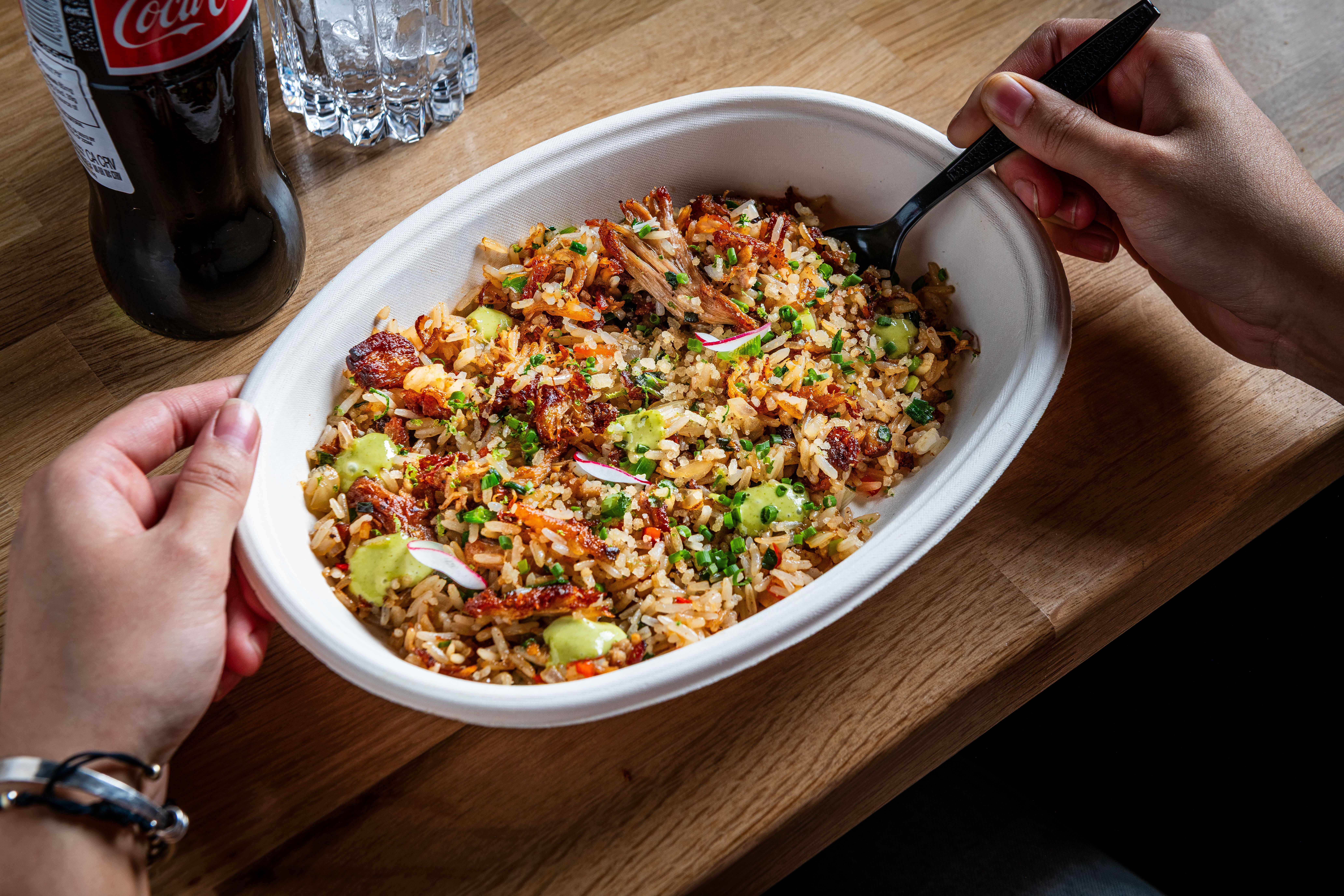 White oval takeout container of fried rice with pork bellu, peas, onions, and a green sauce, held by two hands with a fork on a wooden table; a Coca-Cola bottle and a water bottle are nearby.