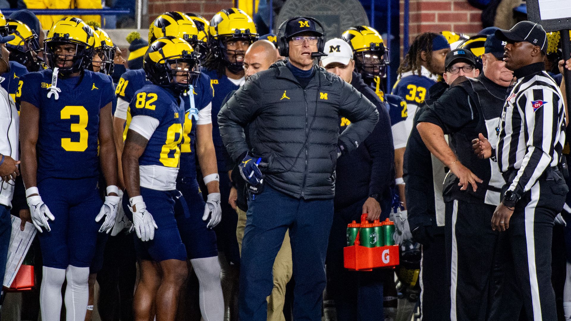 Harbaugh stands in the middle of the frame looking outward with a microphone headset. There are players milling about behind him.