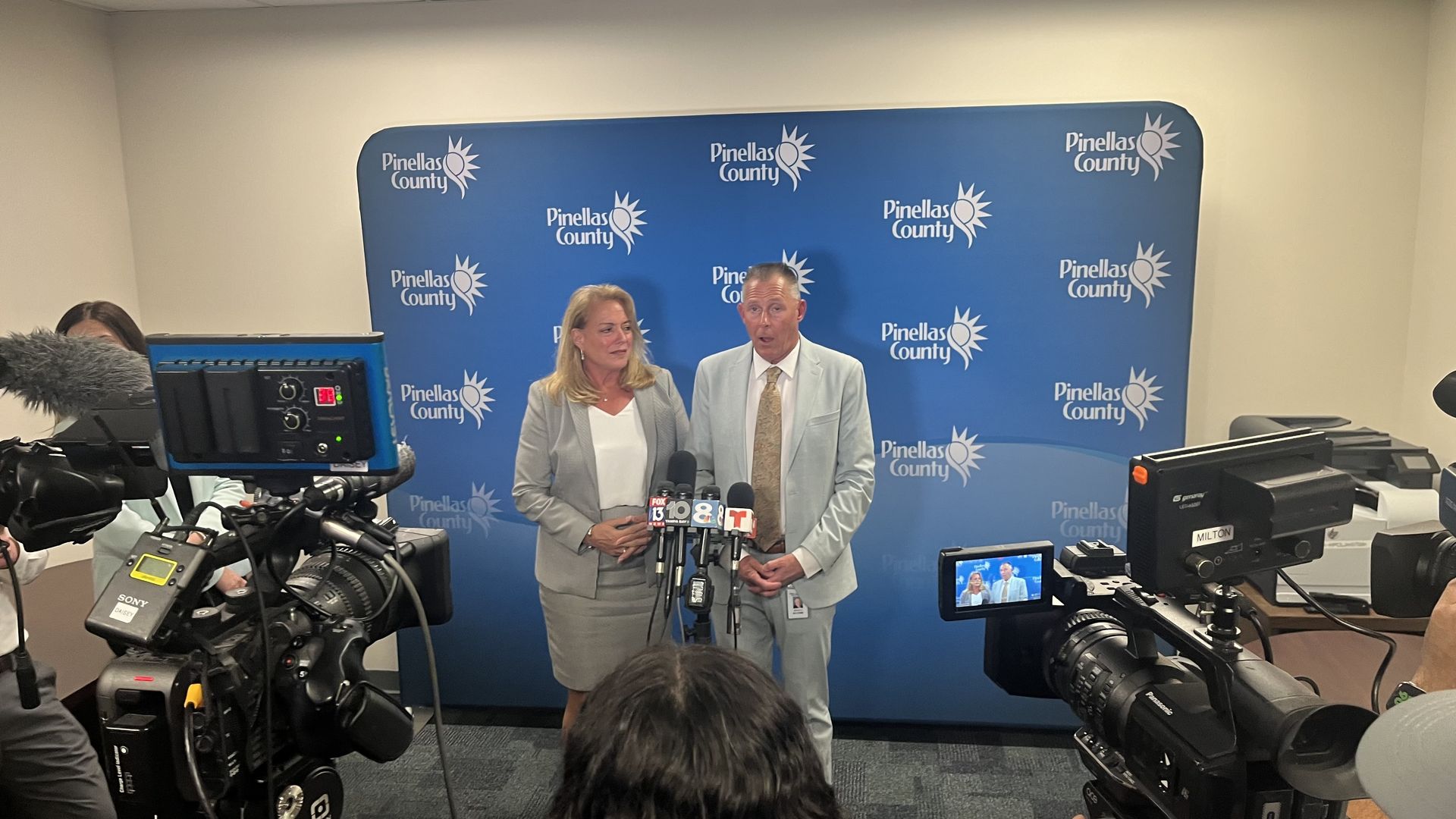 A man and a woman, both wearing gray suits, stand before several TV cameras in front of a blue backdrop dotted with logos that say "Pinellas County."